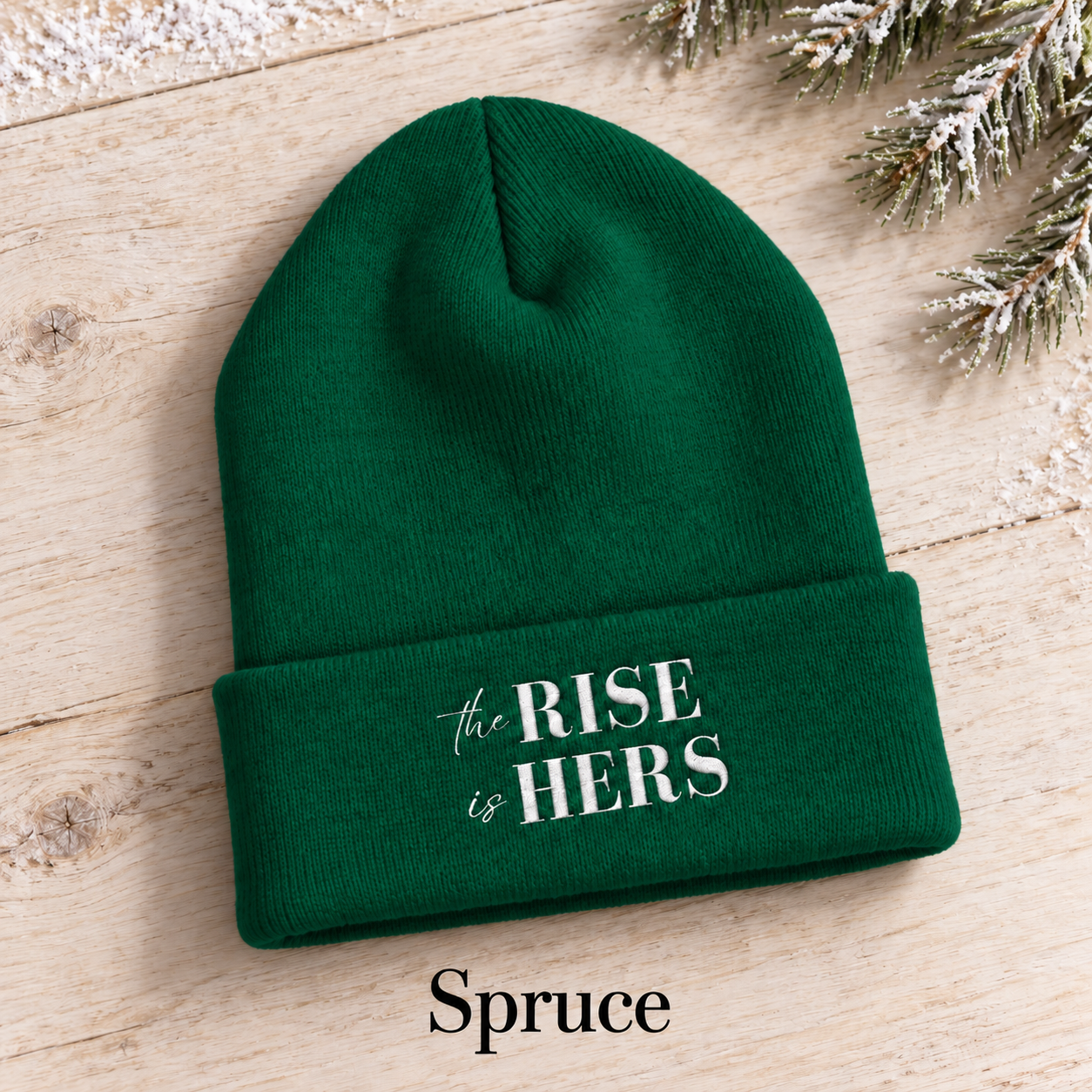 Spruce green The Rise Is Hers cuffed knit beanie with an embroidered logo, displayed on a light background beside an evergreen branch.
