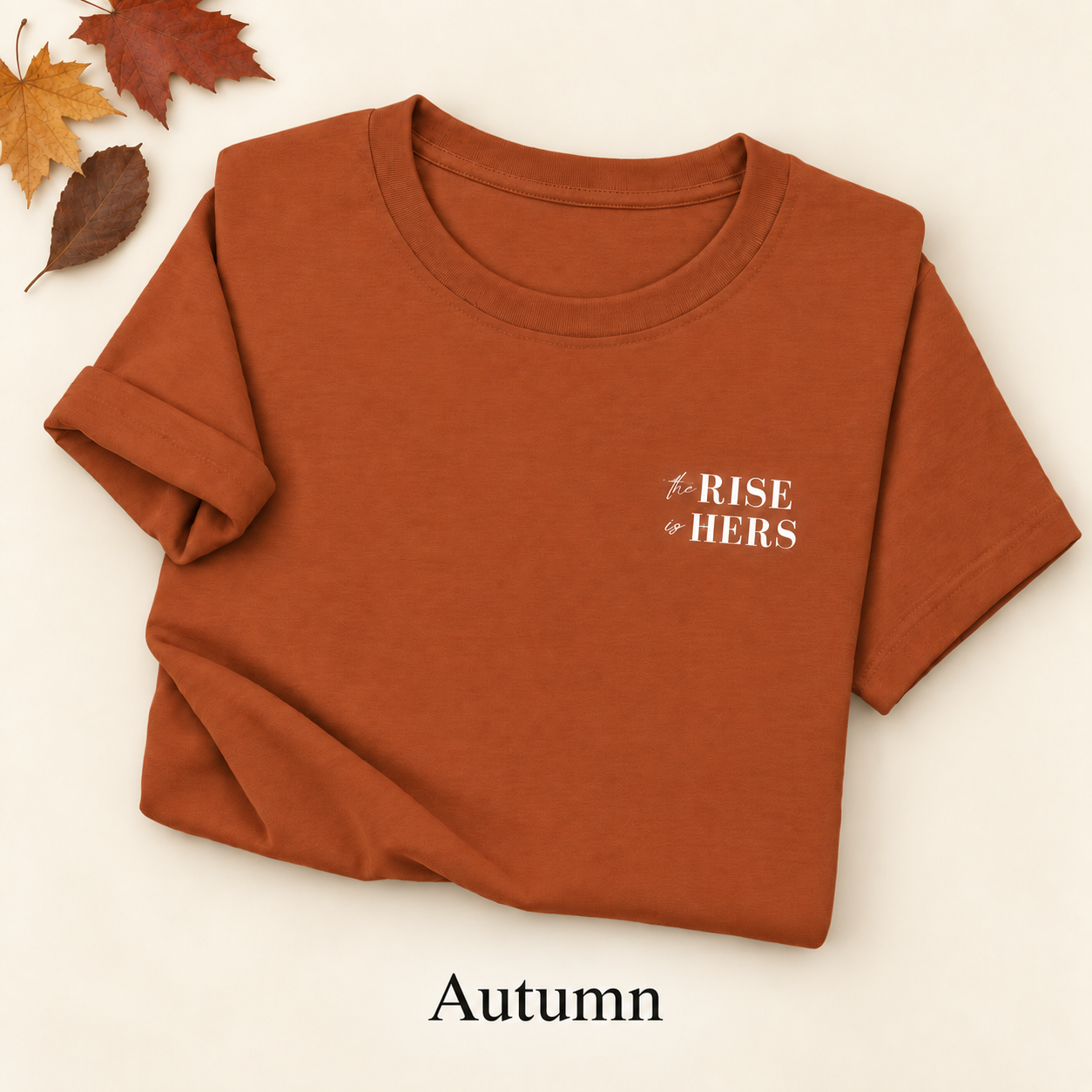 Autumn rust The Rise Is Hers T-shirt with an embroidered left-chest logo, folded on an ivory background with autumn leaves.