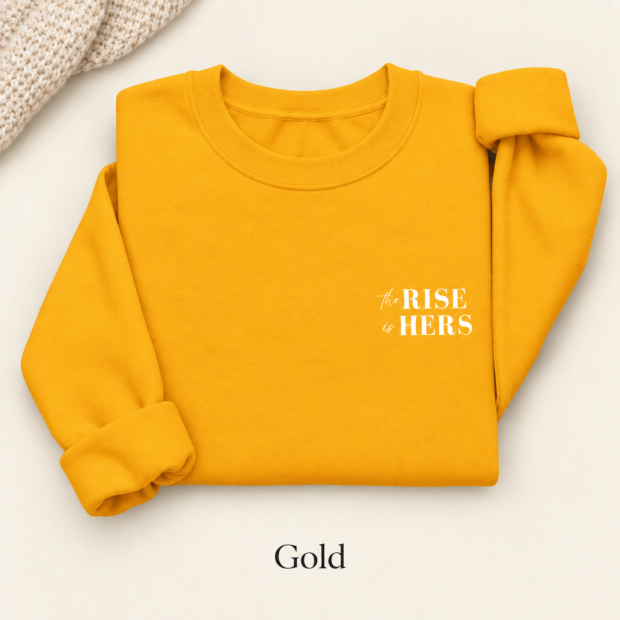 Gold yellow The Rise Is Hers crewneck sweatshirt with an embroidered left-chest logo, folded on an ivory background.