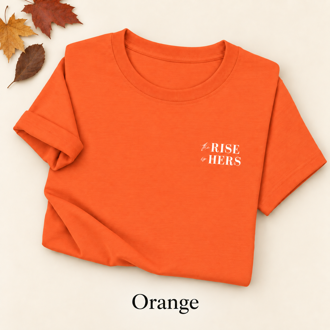 Orange The Rise Is Hers T-shirt with an embroidered left-chest logo, folded on an ivory background with autumn leaves.