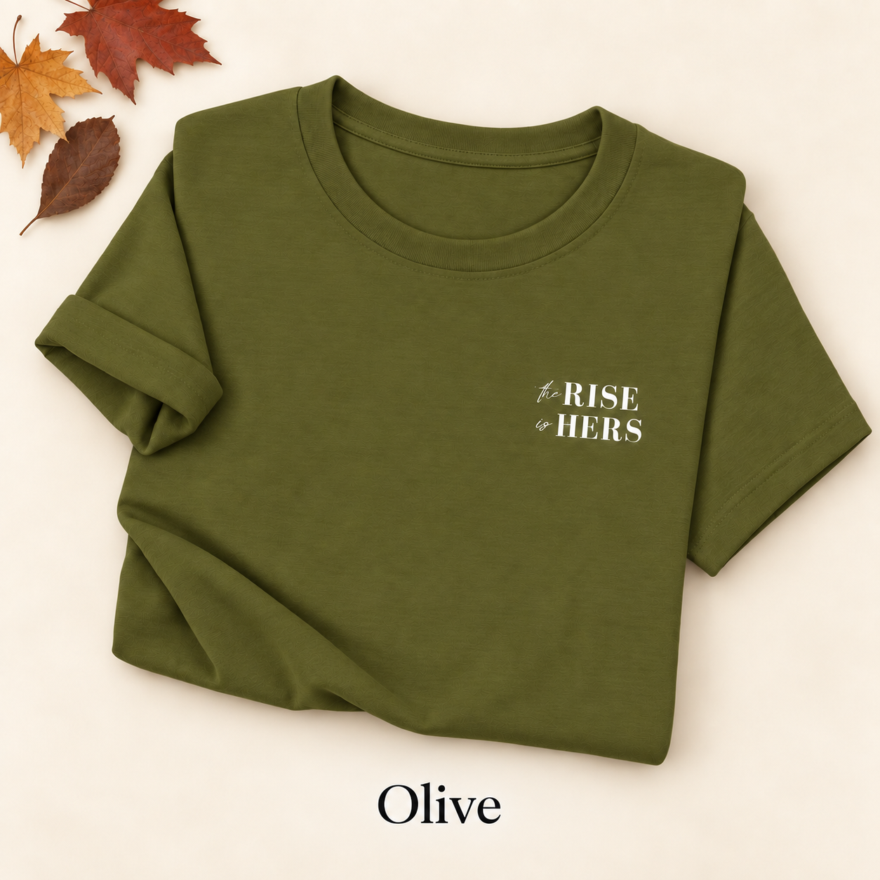Olive green The Rise Is Hers T-shirt with an embroidered left-chest logo, folded on an ivory background with autumn leaves.