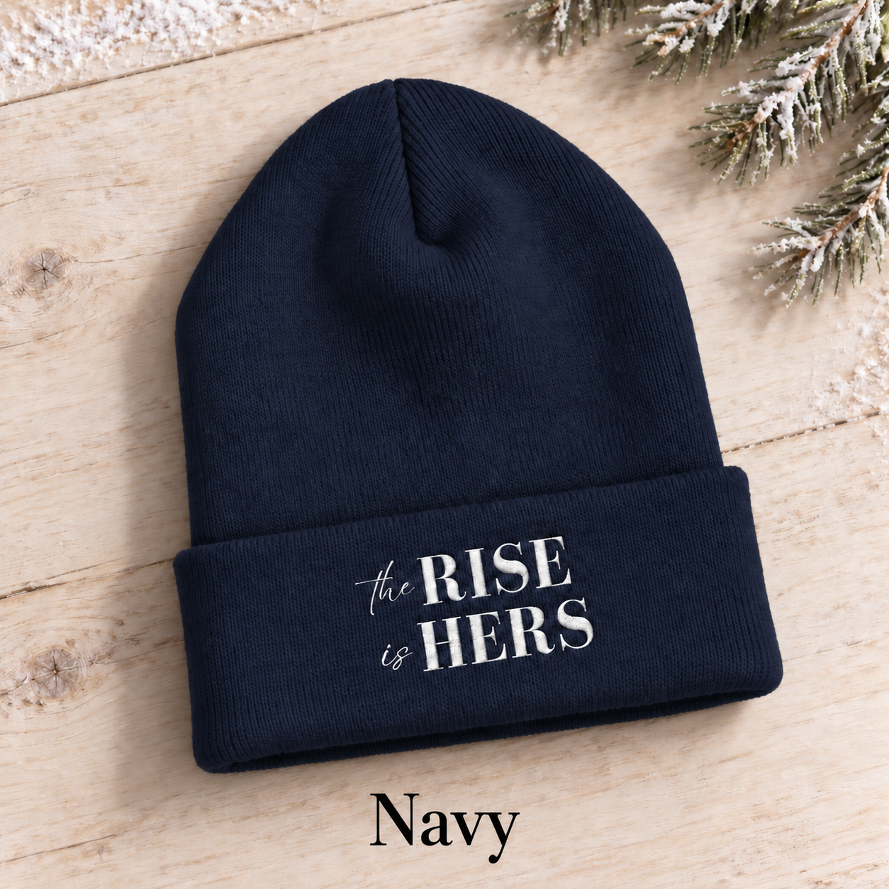 Navy blue The Rise Is Hers cuffed knit beanie with an embroidered logo, displayed on a light background beside an evergreen branch.