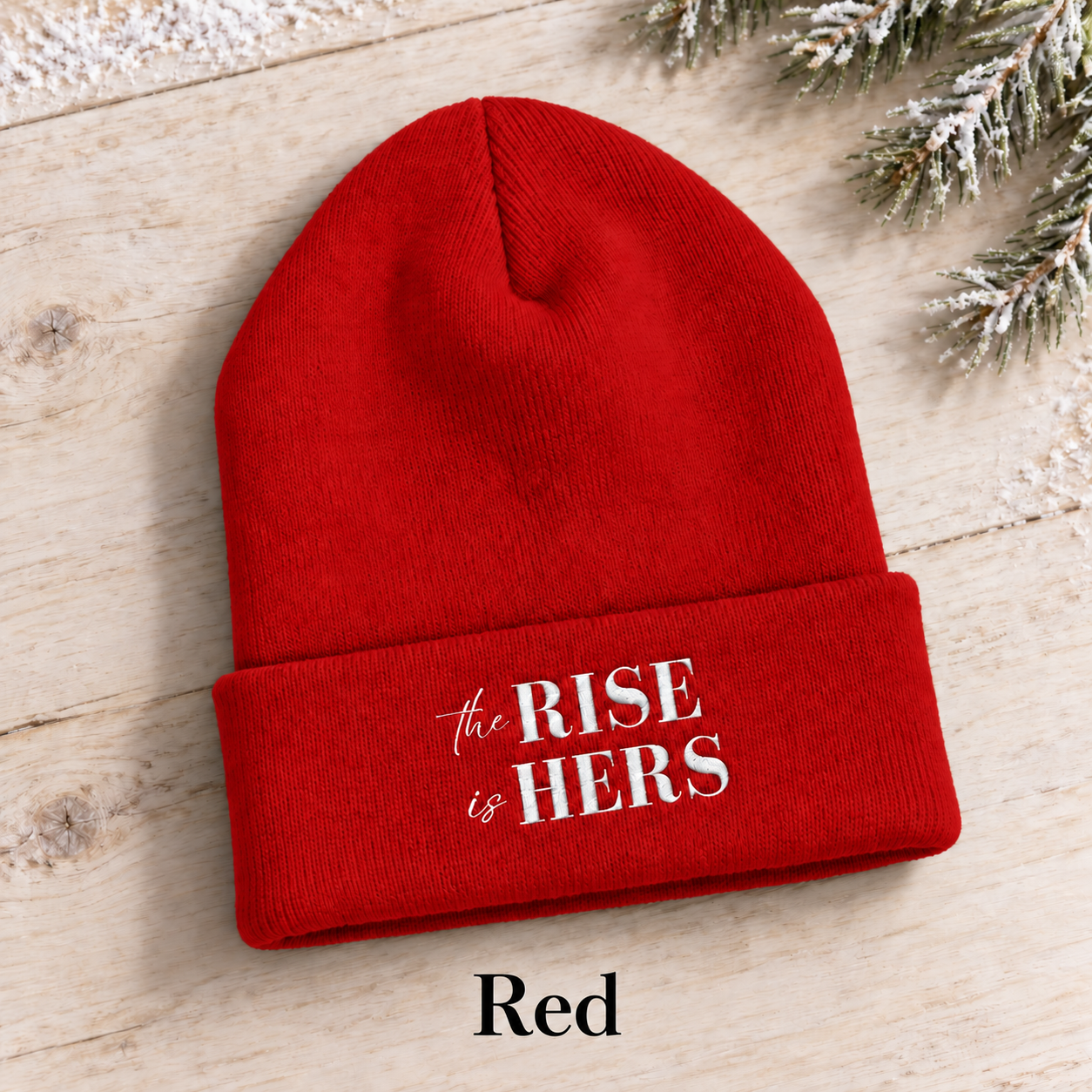 Red The Rise Is Hers cuffed knit beanie with an embroidered logo, displayed on a light background beside an evergreen branch.