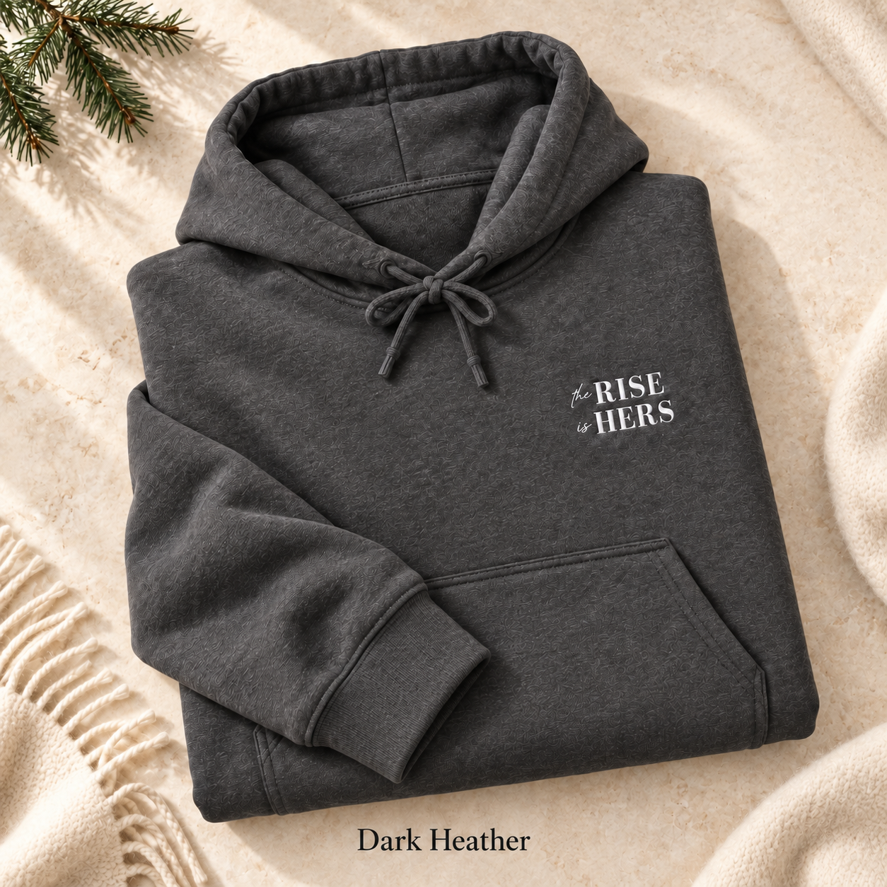 Dark Heather The Rise Is Hers hoodie with an embroidered left-chest logo, folded on cream fabric.