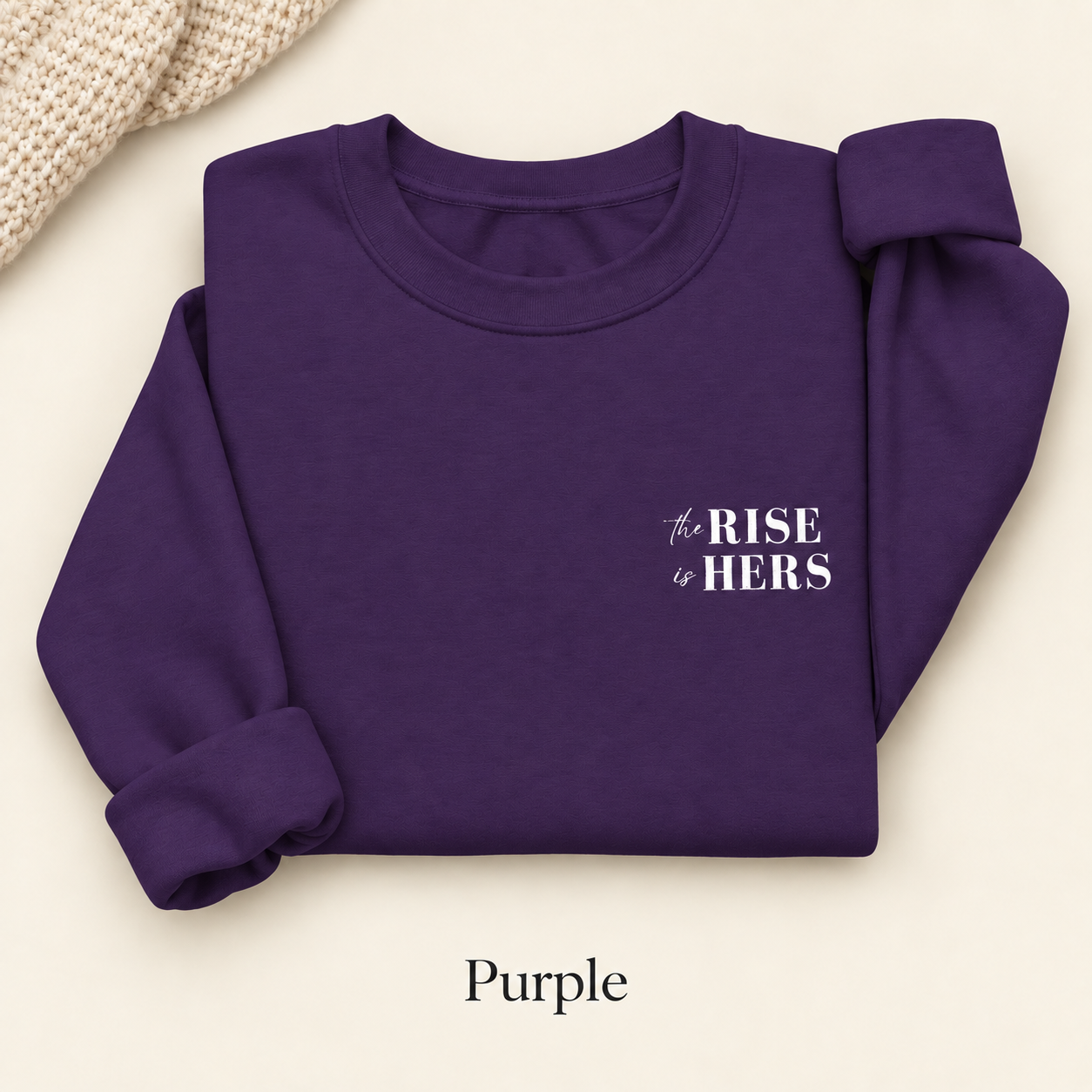 Purple The Rise Is Hers crewneck sweatshirt with an embroidered left-chest logo, folded on an ivory background.
