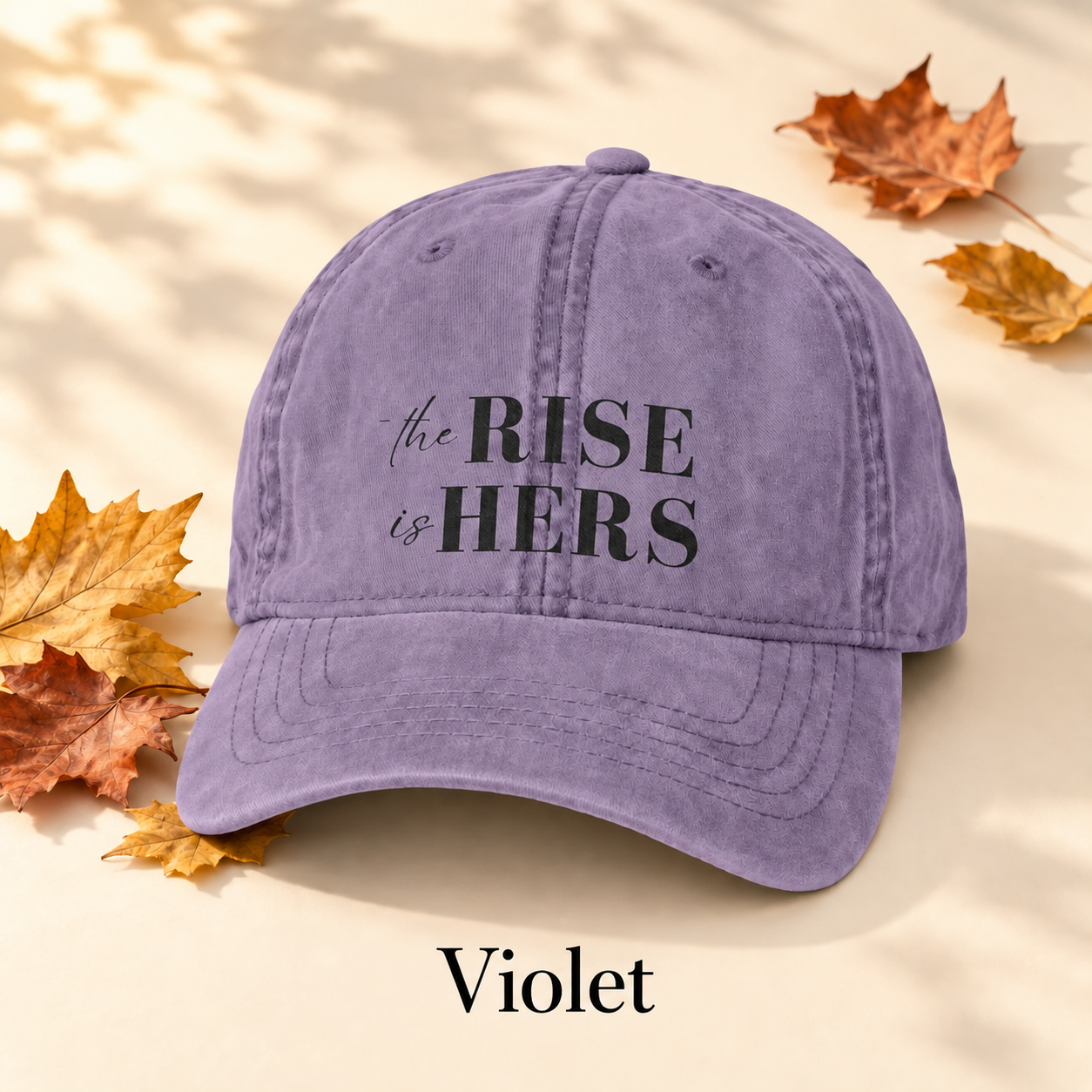Violet The Rise Is Hers baseball hat with an embroidered front logo, displayed beside autumn leaves.