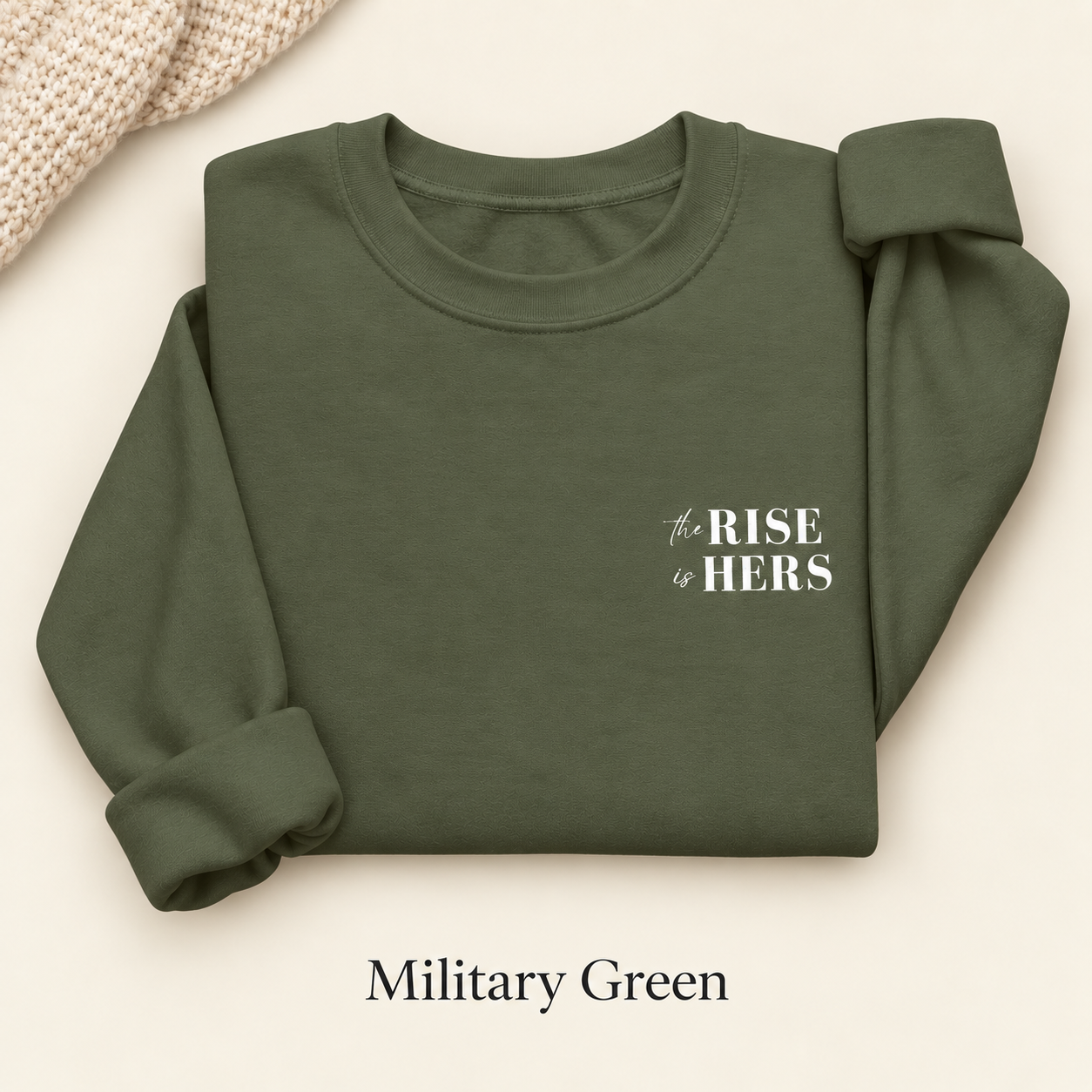 Military Green The Rise Is Hers crewneck sweatshirt with an embroidered left-chest logo, folded on an ivory background.