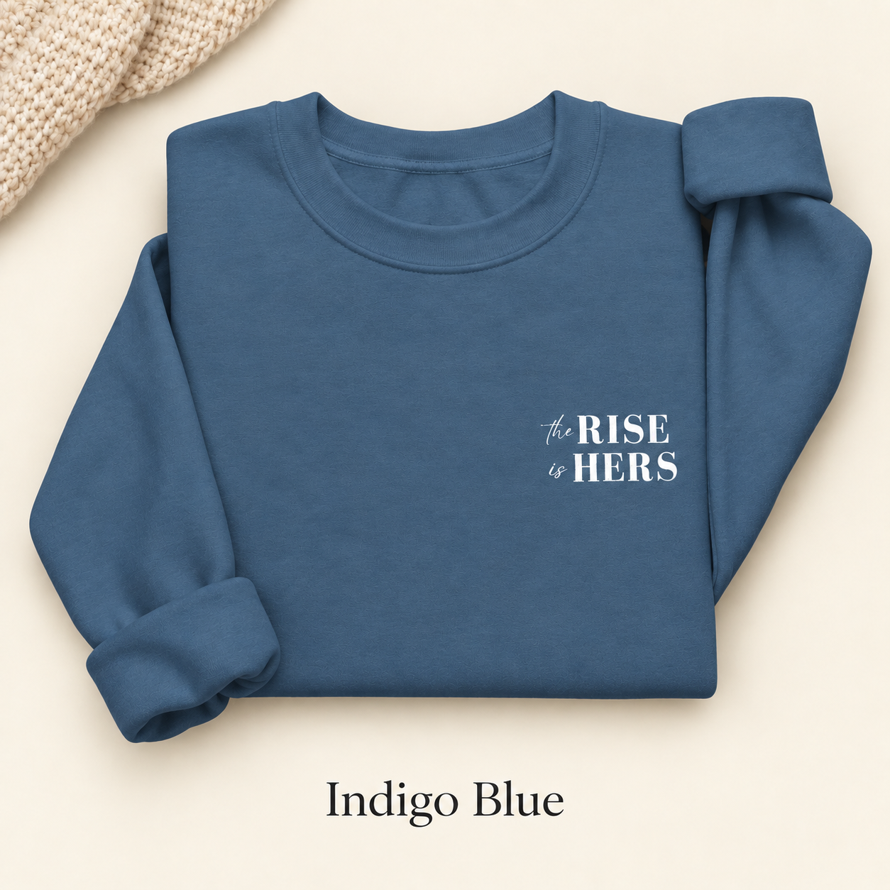 Indigo Blue The Rise Is Hers crewneck sweatshirt with an embroidered left-chest logo, folded on an ivory background.