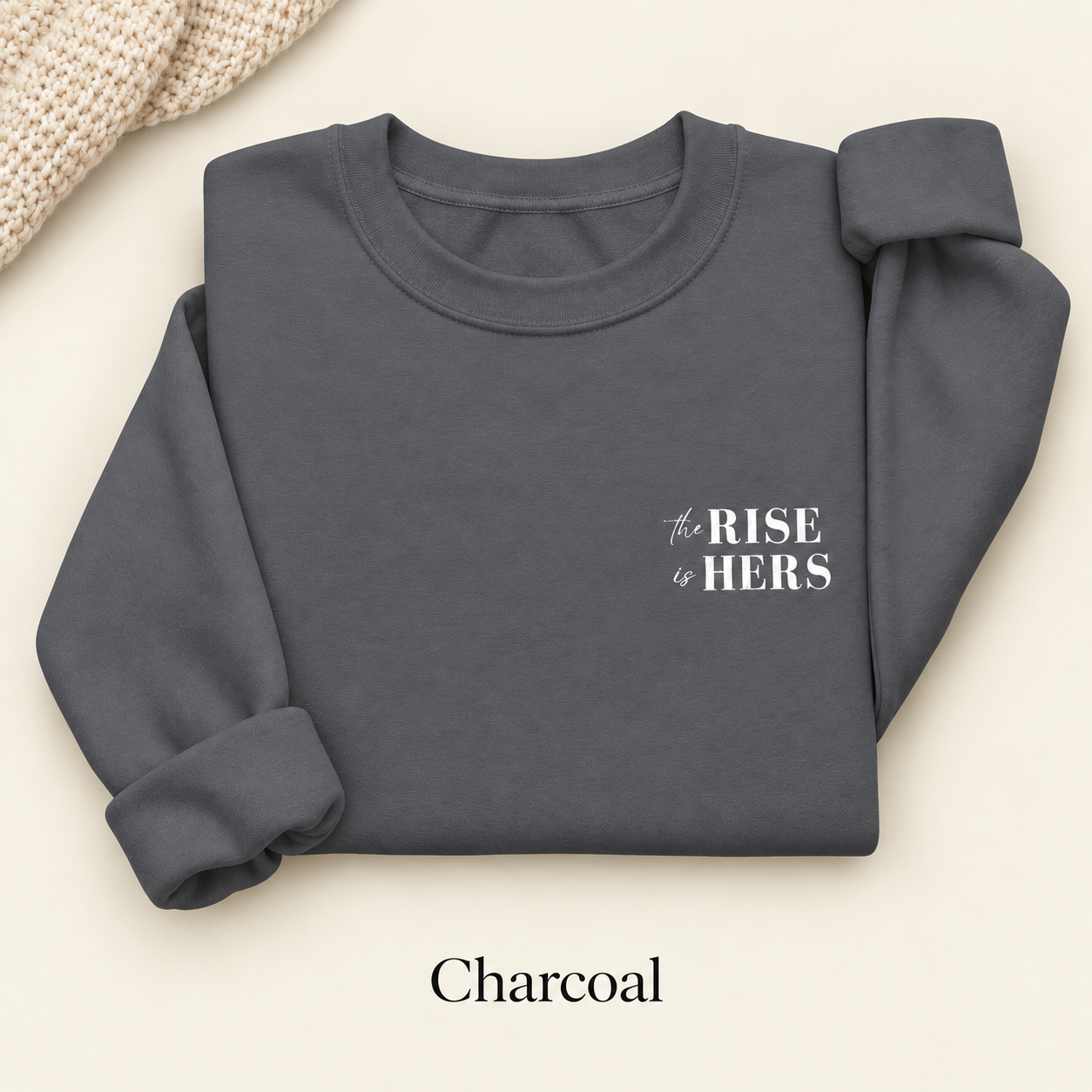 Charcoal gray The Rise Is Hers crewneck sweatshirt with an embroidered left-chest logo, folded on an ivory background.