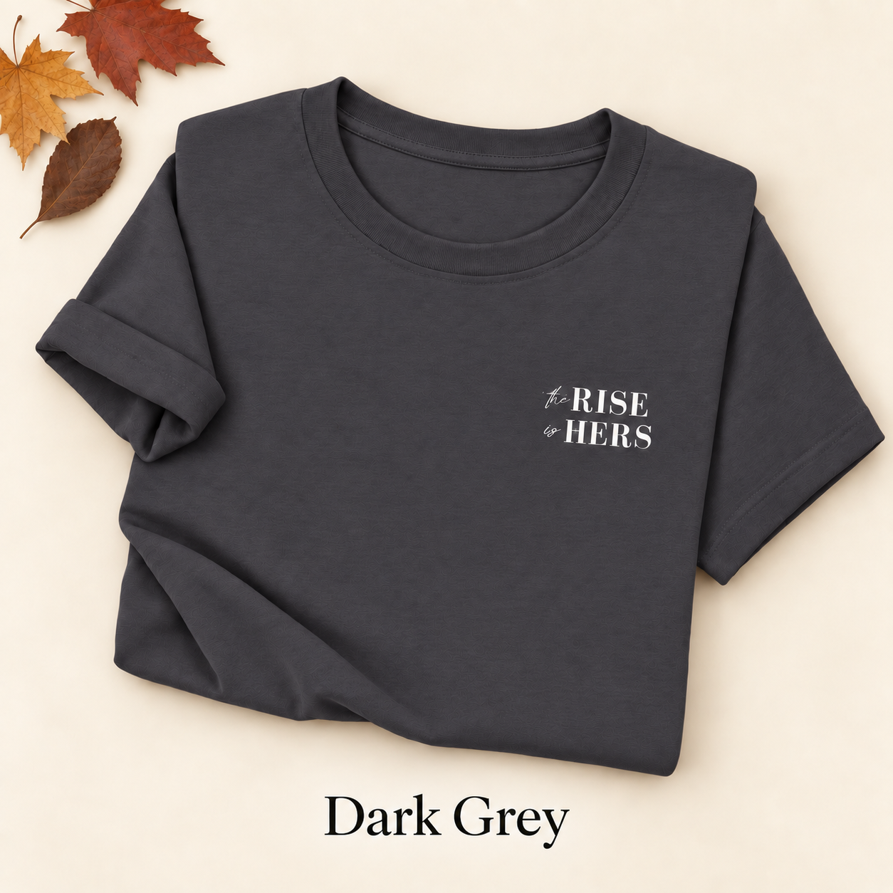 Dark Grey The Rise Is Hers T-shirt with an embroidered left-chest logo, folded on an ivory background with autumn leaves.