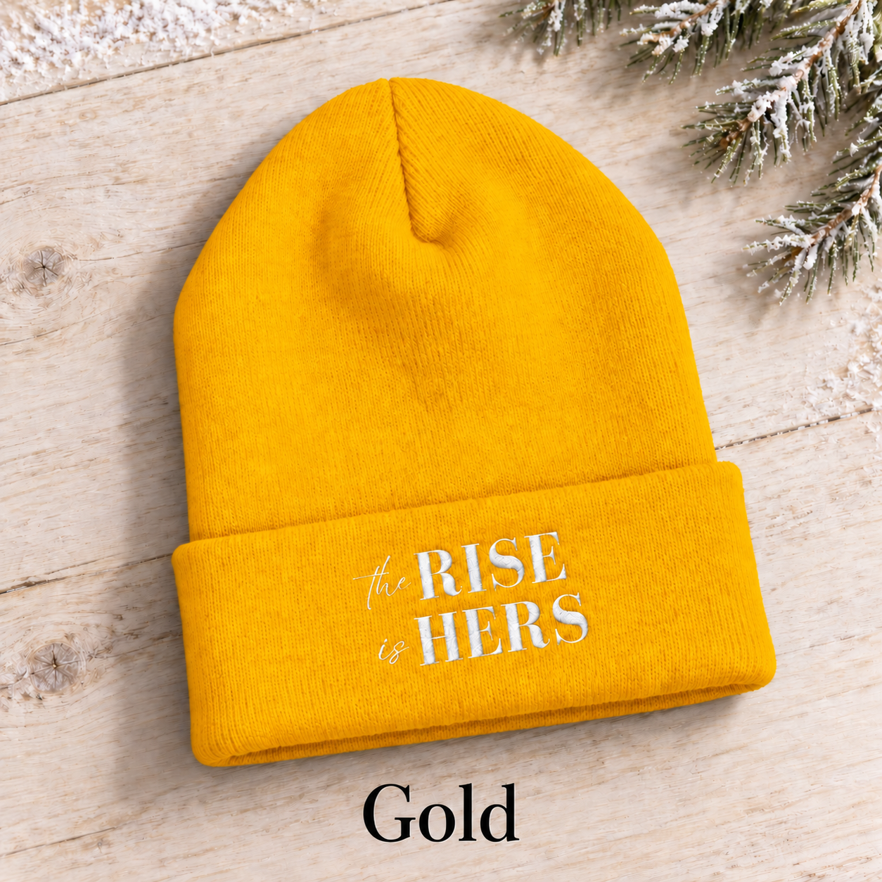 Gold yellow The Rise Is Hers cuffed knit beanie with an embroidered logo, displayed on a light background beside an evergreen branch.