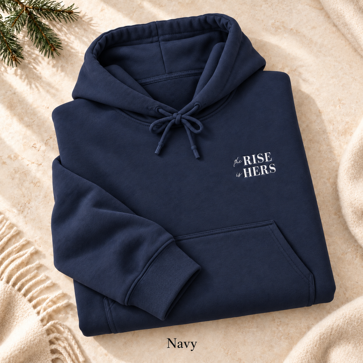 Navy The Rise Is Hers hoodie with an embroidered left-chest logo, folded on cream fabric.