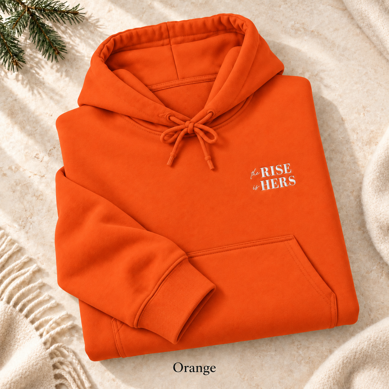 Orange The Rise Is Hers hoodie with an embroidered left-chest logo, folded on cream fabric.