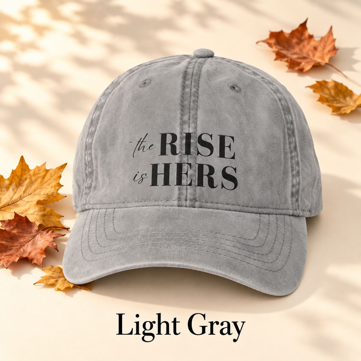 Light Gray The Rise Is Hers baseball hat with an embroidered front logo, displayed beside autumn leaves.