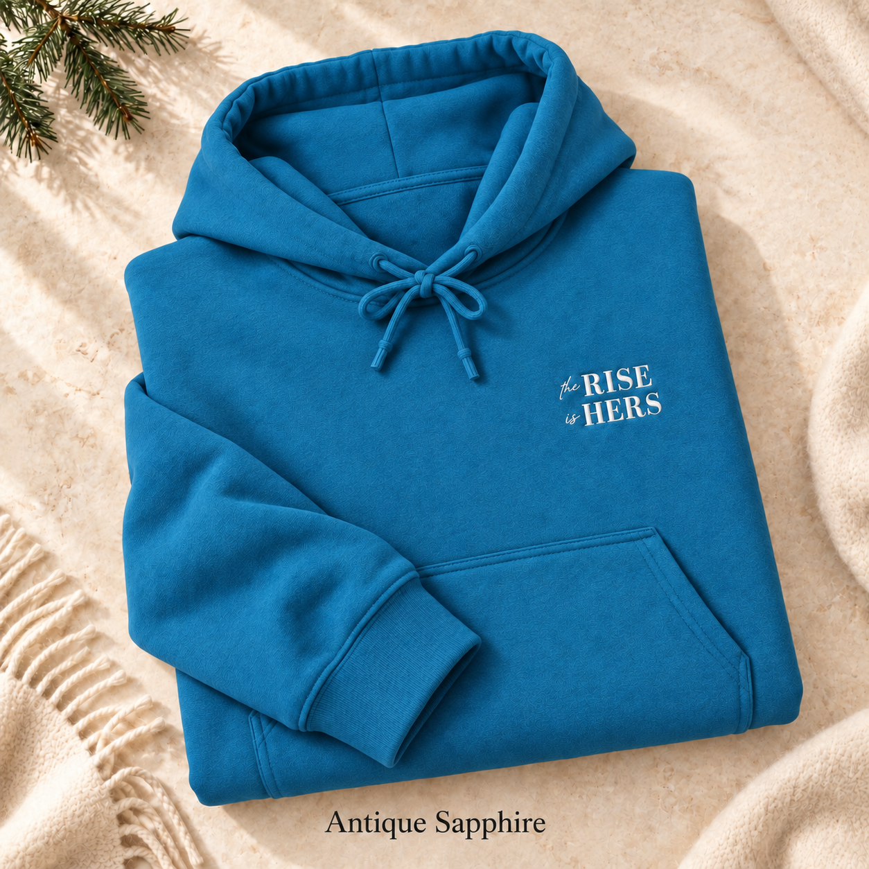 Antique Sapphire The Rise Is Hers hoodie with an embroidered left-chest logo, folded on cream fabric.