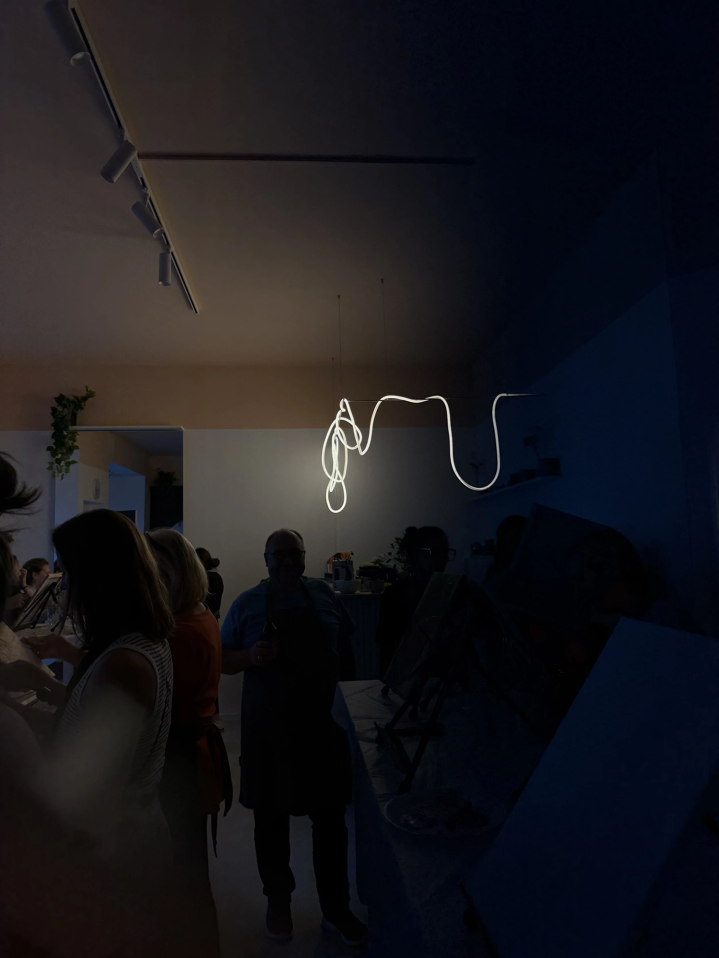 People gathered in a dimly lit room with a modern, white, abstract light fixture hanging from the ceiling, and a small plant on a shelf in the background.