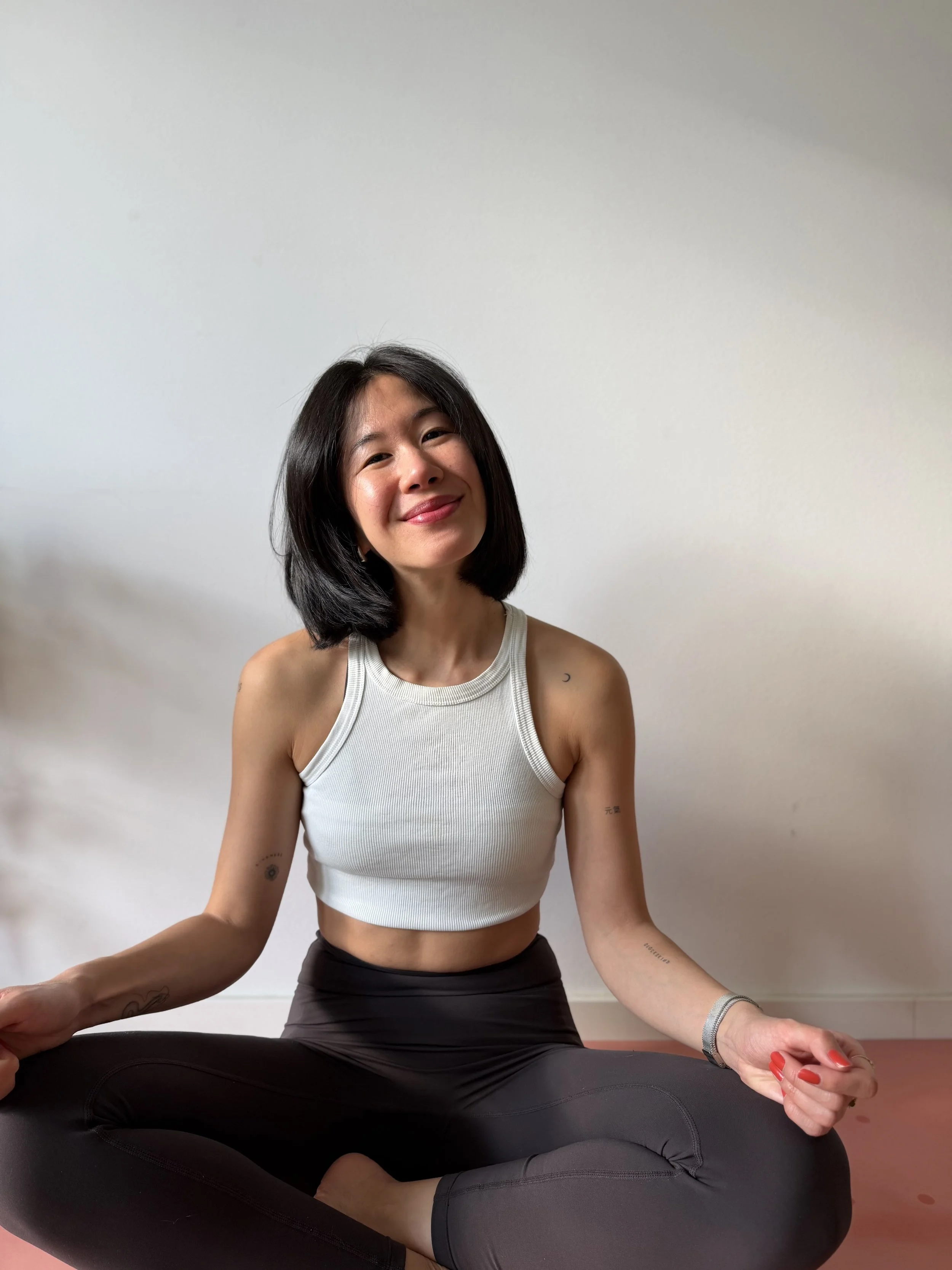 A woman practicing yoga in a cross-legged seated position, sitting on a yoga mat, with her hands resting on her knees and fingers in a mudra. She is smiling, wearing a sleeveless white athletic top and dark leggings, with a plain white wall in the background.