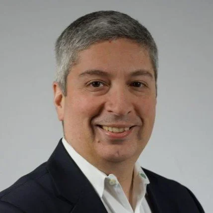 Portrait of a man with short gray hair, wearing a white collared shirt and dark blazer, smiling against a plain gray background.