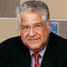 A close-up portrait of an older man with gray hair, glasses, wearing a dark suit and red tie, smiling at the camera.