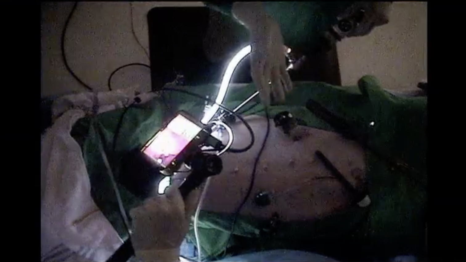 Person using a smartphone and a makeshift electronic device in a dimly lit room with a bed and random tools.