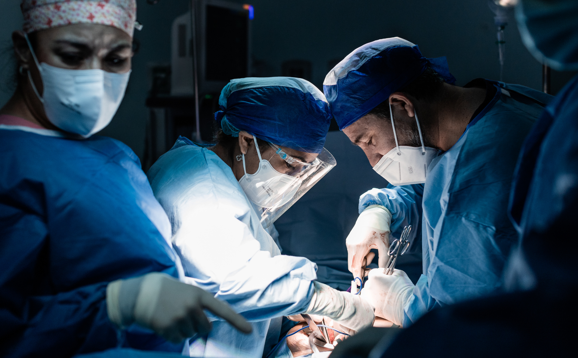 Medical team performing surgery in an operating room, wearing scrubs, caps, masks, and protective eyewear, focused on the procedure.