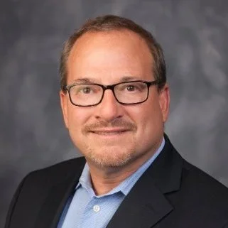 Portrait of a middle-aged man with glasses, wearing a suit jacket and light blue shirt, smiling against a gray studio background.