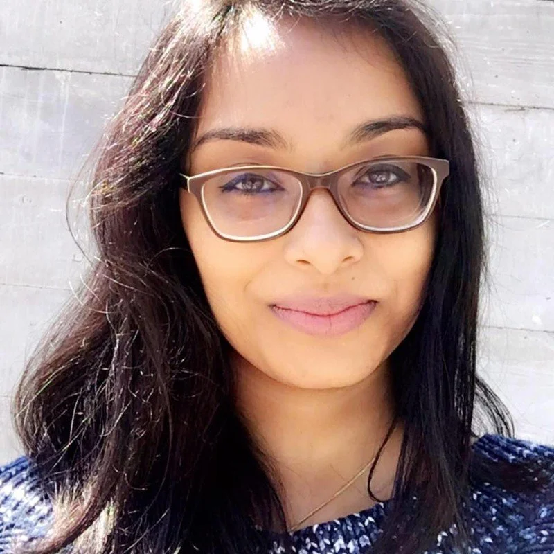 Close-up of a woman with long dark hair, wearing glasses and a blue patterned top, smiling outdoors with sunlight and a light gray wall in the background.