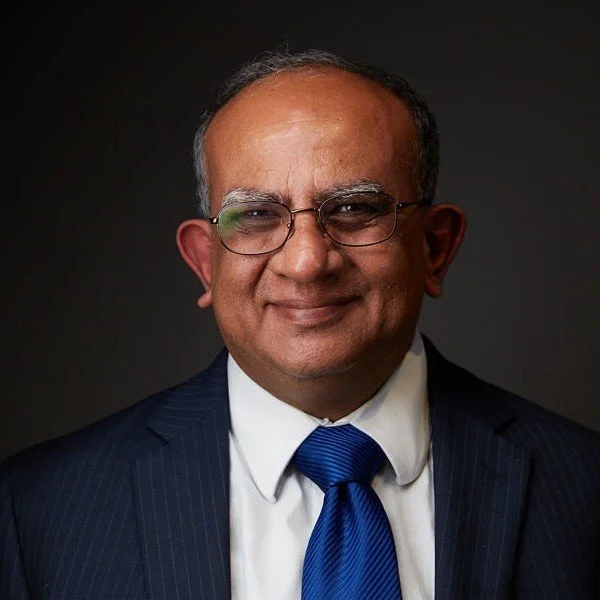 A professional portrait of a middle-aged man wearing glasses, a dark suit with pinstripes, a white shirt, and a blue tie, smiling against a dark background.