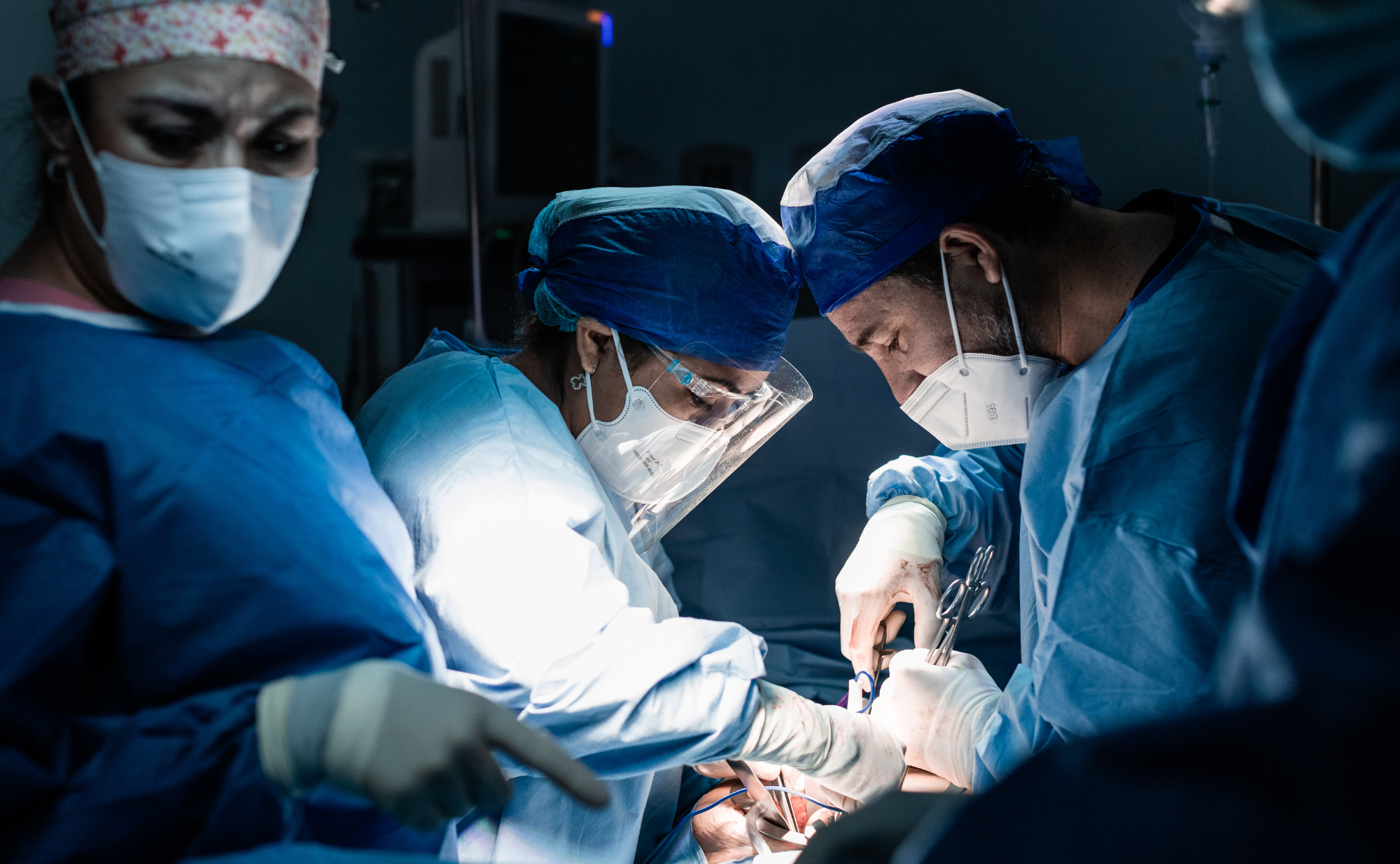 A team of surgeons performing surgery in an operating room, wearing blue surgical gowns, masks, and head coverings, focused on the procedure.