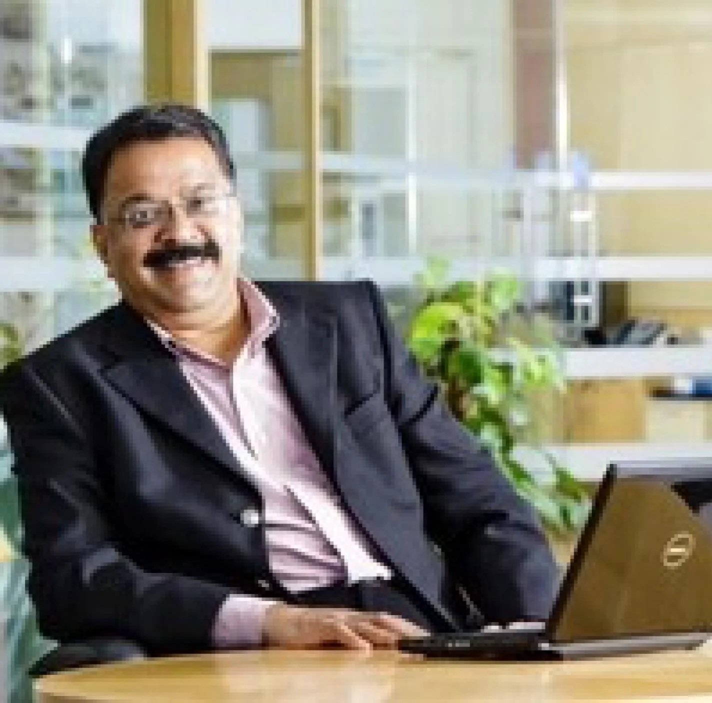 A man with dark hair, glasses, and a mustache, wearing a black suit and pink shirt, sitting at a desk with a laptop, smiling in an office with glass walls and green plants.