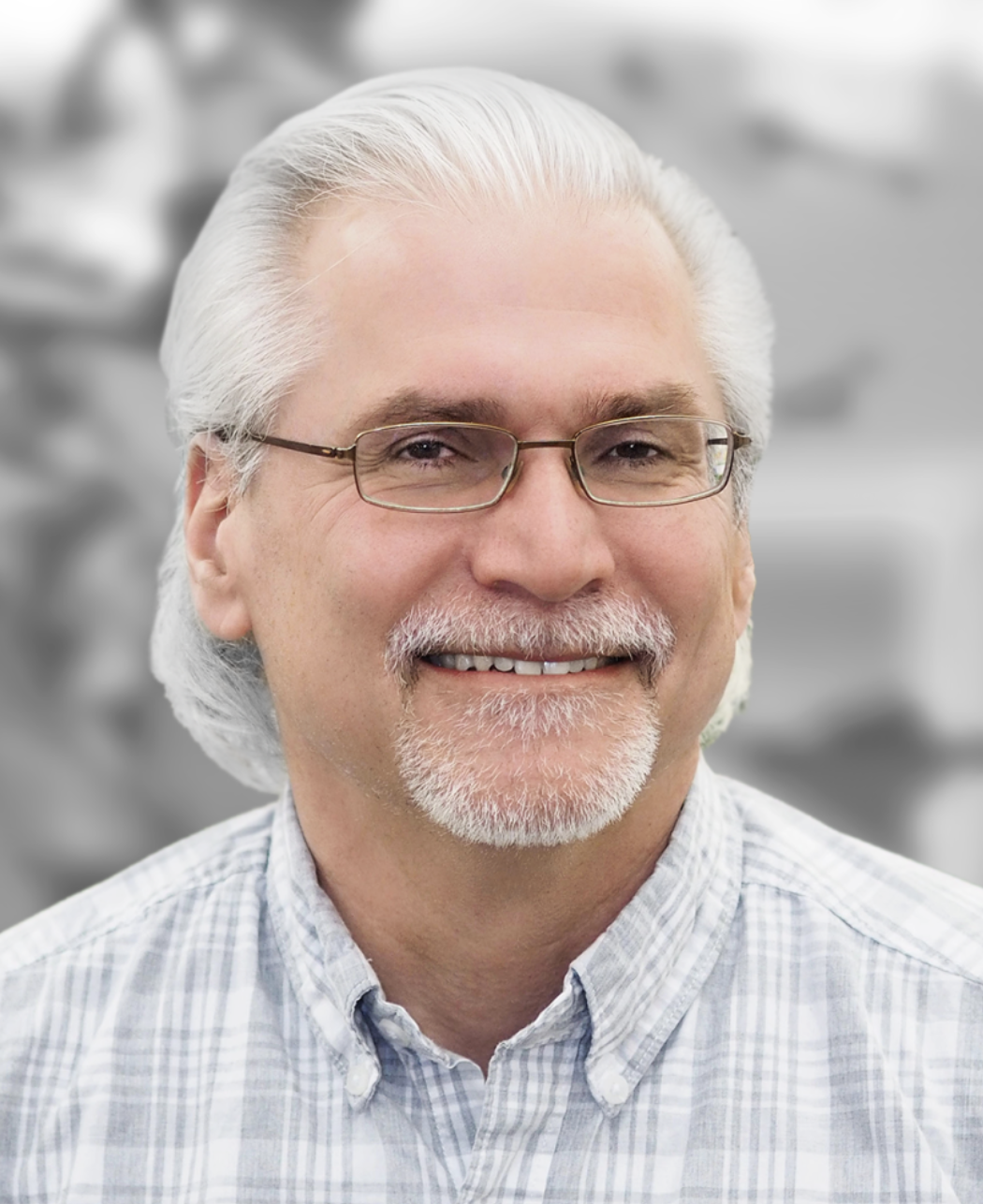 A smiling middle-aged man with white hair, glasses, and a beard, wearing a light blue checkered shirt, indoors with a blurred background.