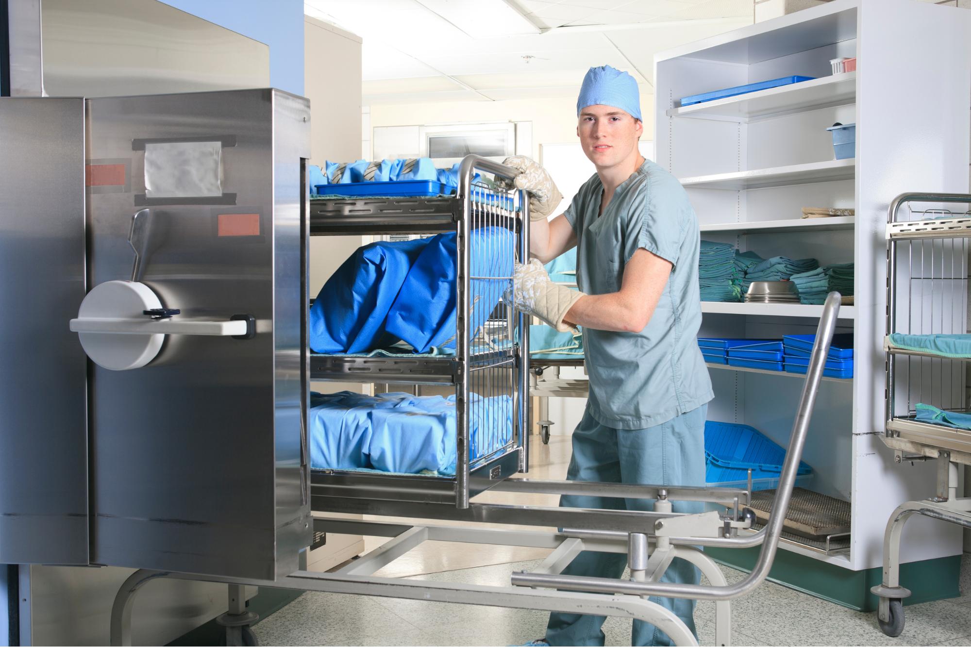 A young male healthcare worker in scrubs and a blue cap is moving a cart loaded with blue hospital linen in a medical facility's storage room.