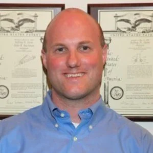 A smiling man with a shaved head wearing a light blue collared shirt, standing in front of framed certificates or diplomas.