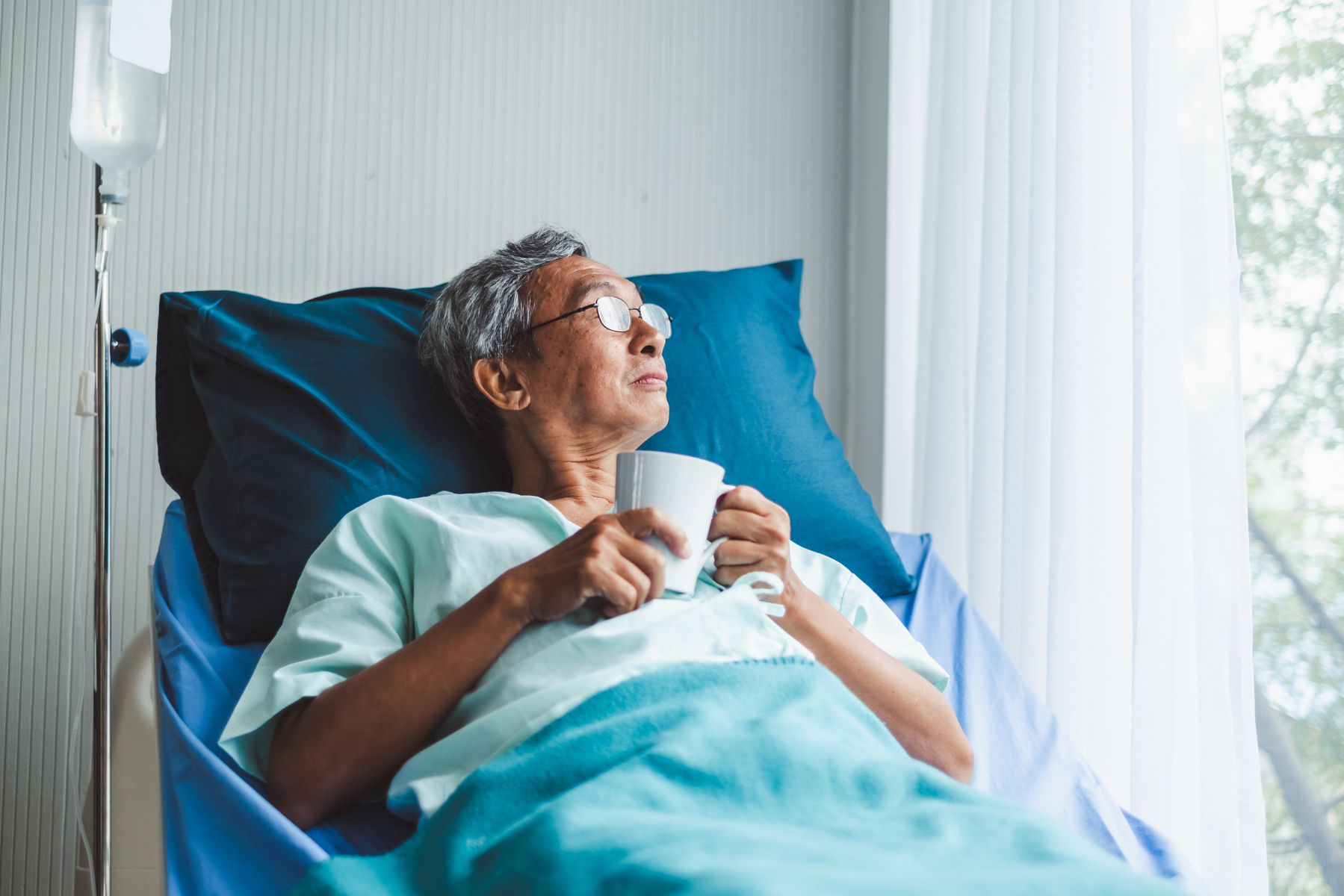 An elderly woman in a hospital bed, wearing glasses, holding a white mug, gazing out the window.
