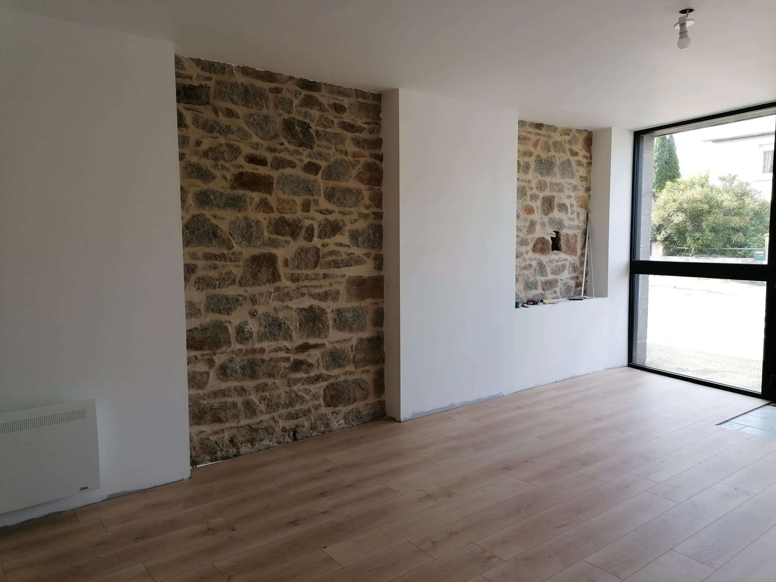 Intérieur d'une pièce avec un mur en pierre apparente, un sol en bois clair, une grande fenêtre laissant entrer la lumière naturelle.