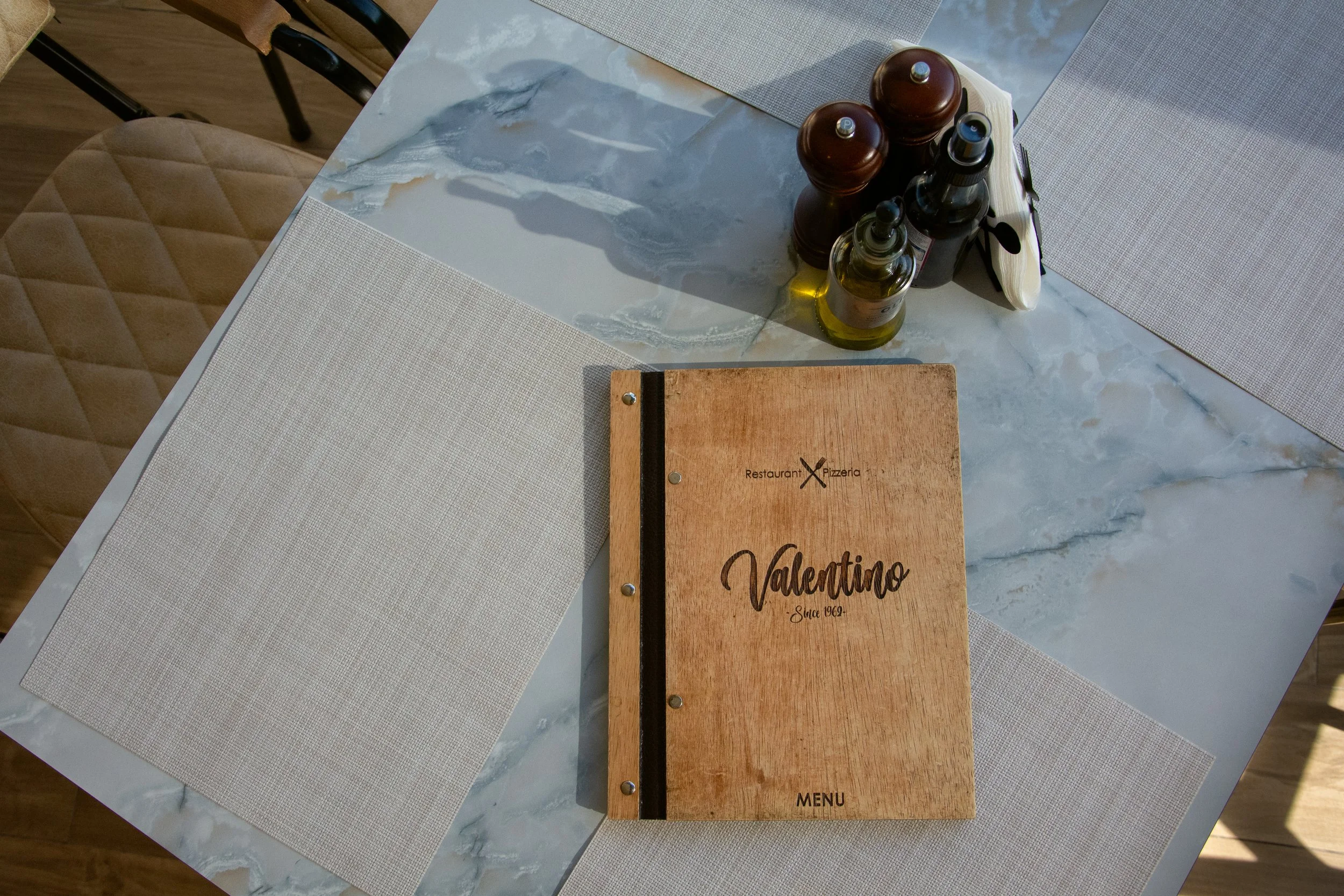 A marble dining table with place mats, a menu labeled 'Valentino,' and a set of salt and pepper shakers, olive oil, and pepper spray on top.