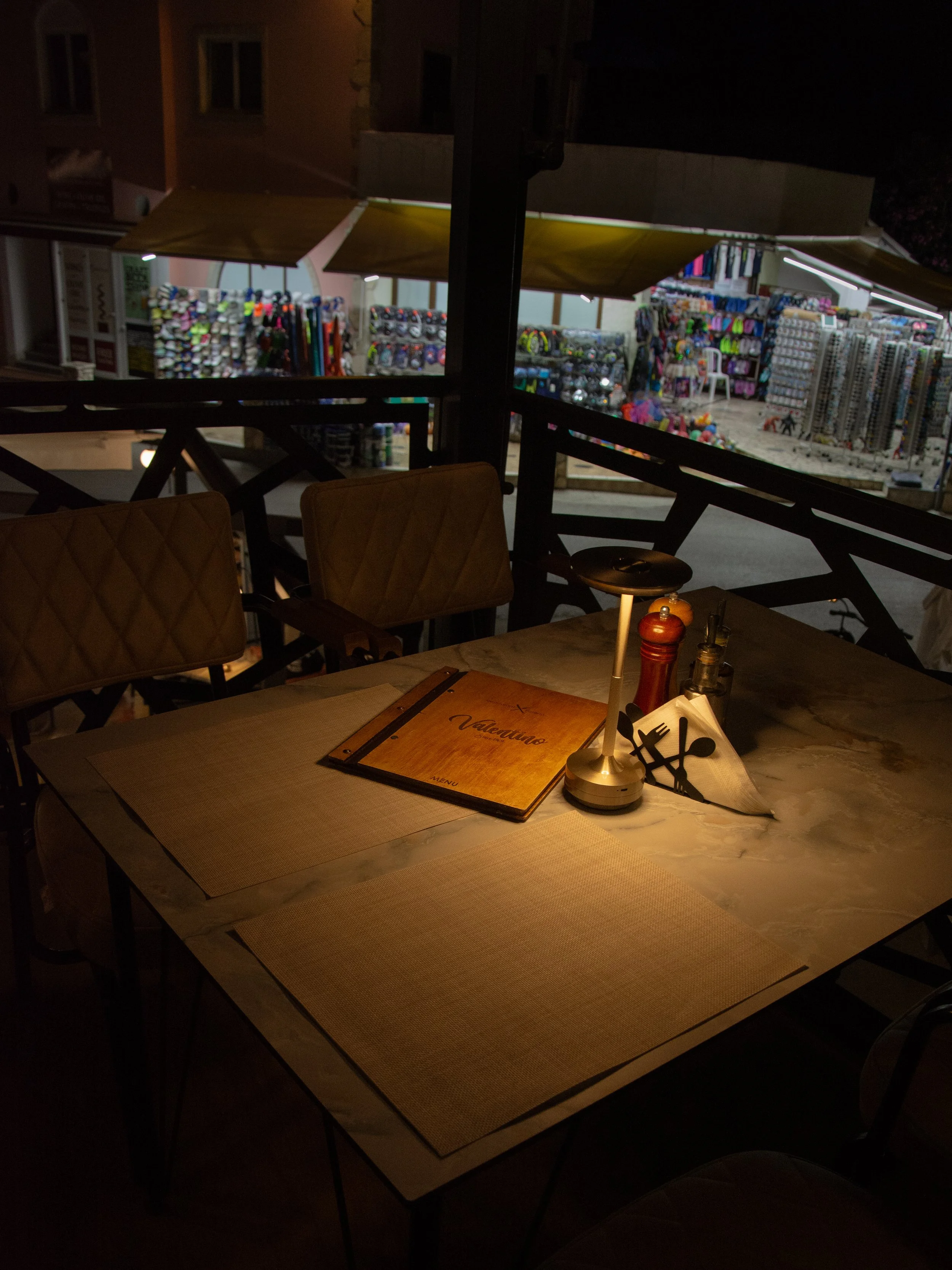A table at a restaurant with a menu, salt and pepper shakers, a small lamp, and napkins. The view outside shows a brightly lit store selling various colorful accessories and toys at night.