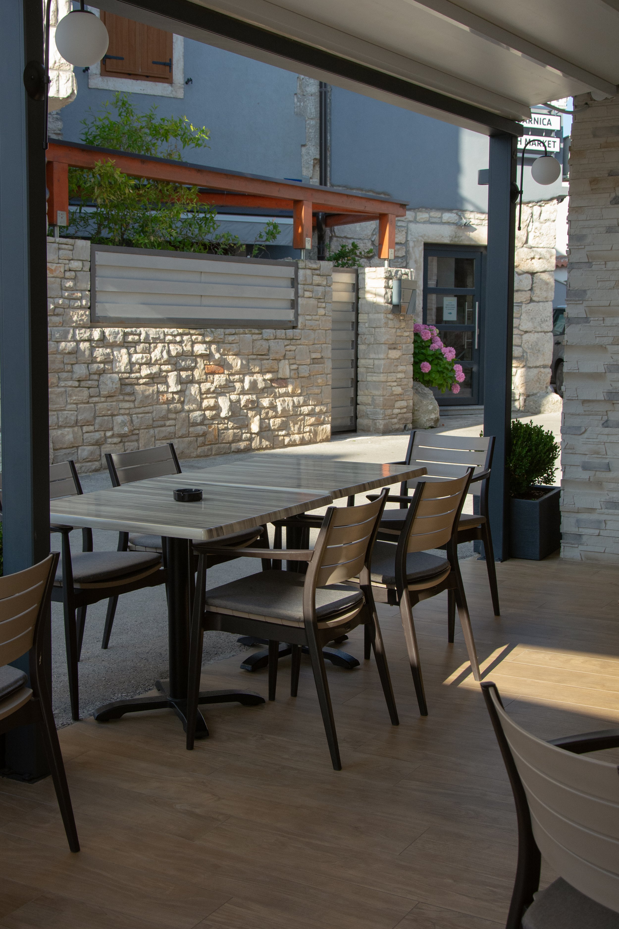 Outdoor seating area with a wooden table and metal chairs, partially shaded, with a view of stone and brick buildings, pink flowering plants, and a blue building wall in the background.