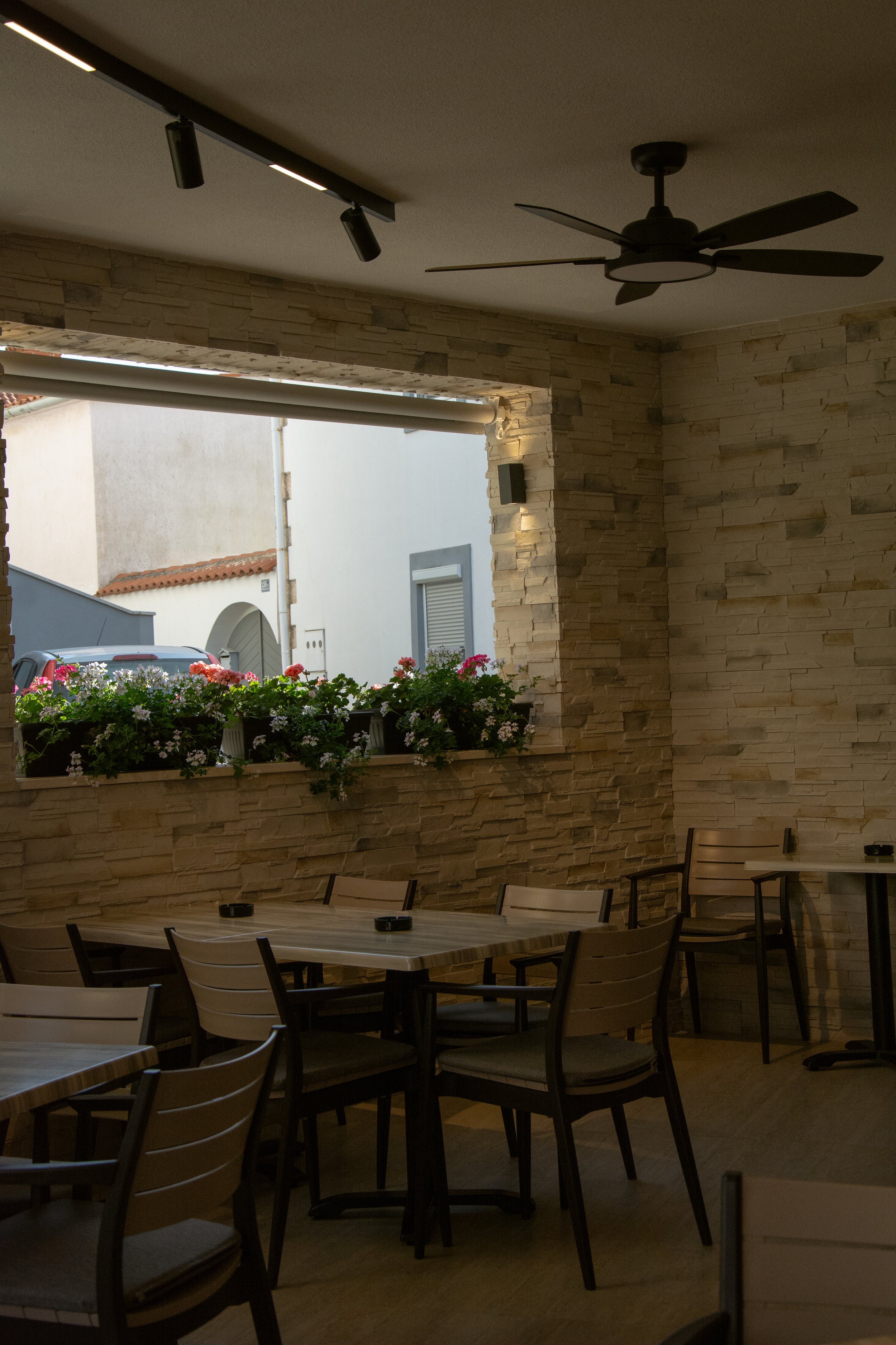 An outdoor patio area with wooden tables and chairs, a brick wall, hanging flower pots with pink and white flowers, and a large open window showing neighboring buildings with white walls and red-tiled roofs.