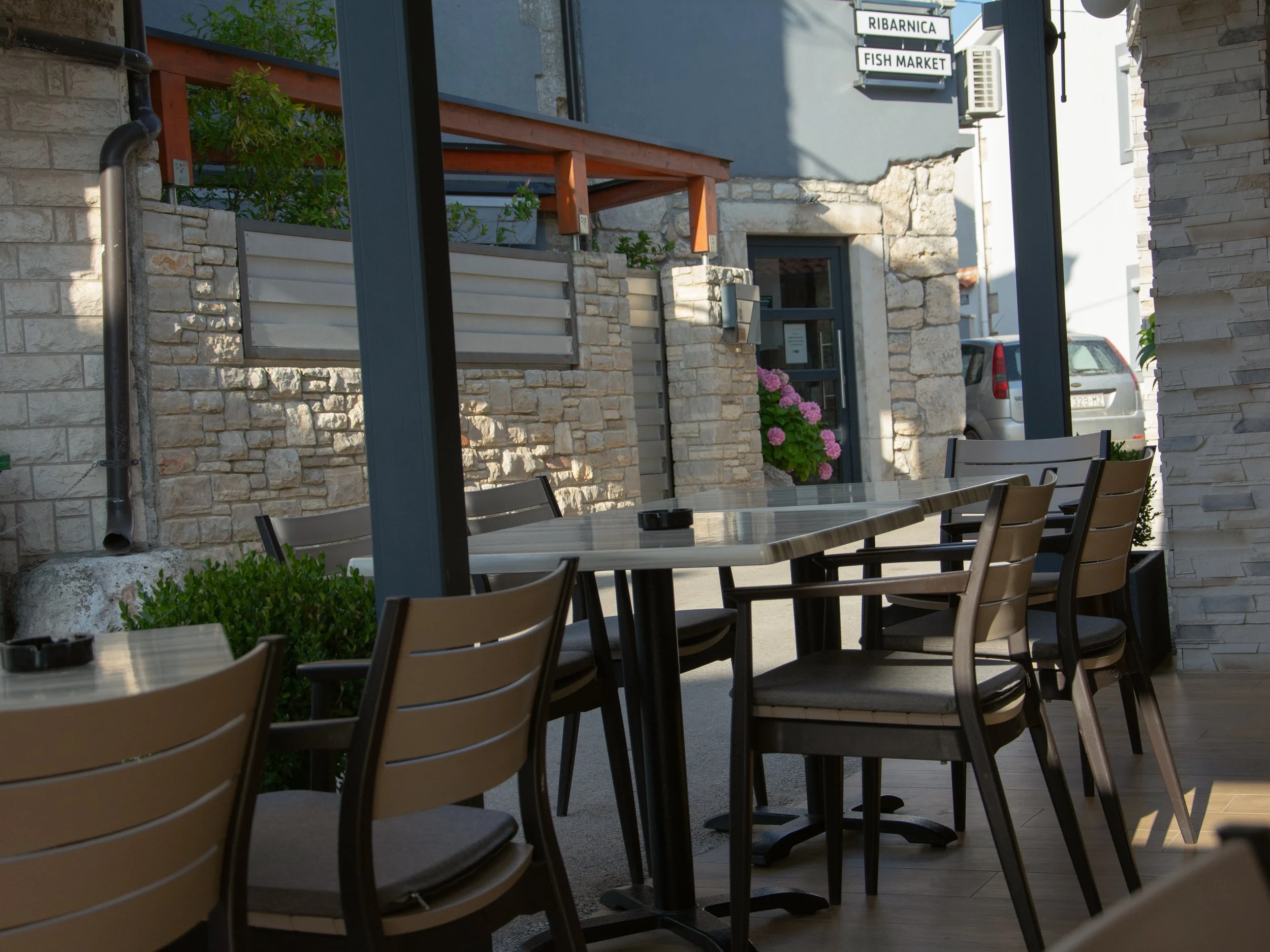 Outdoor seating area with wooden tables and chairs, stone walls, potted pink flowers, and signs for 'Ribarnica' and 'Fish Market' on the building in the background.
