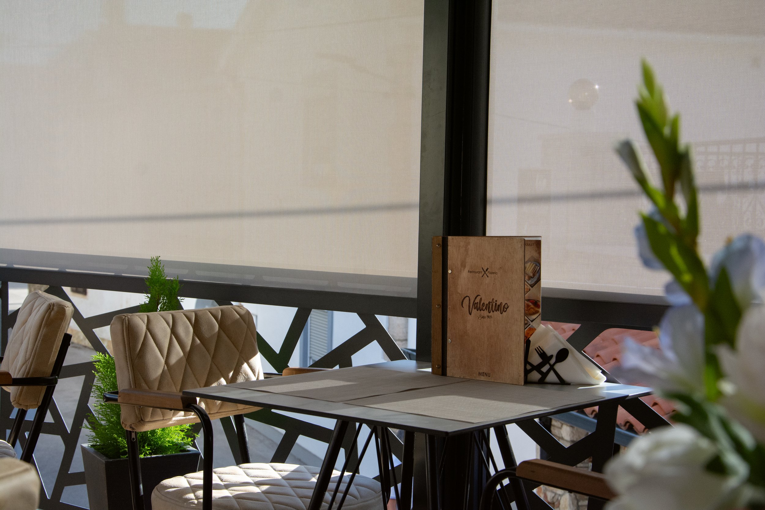 Indoor restaurant setting with beige cushioned chairs, a black metal table, and potted plants, with a large window and a menu on the table.