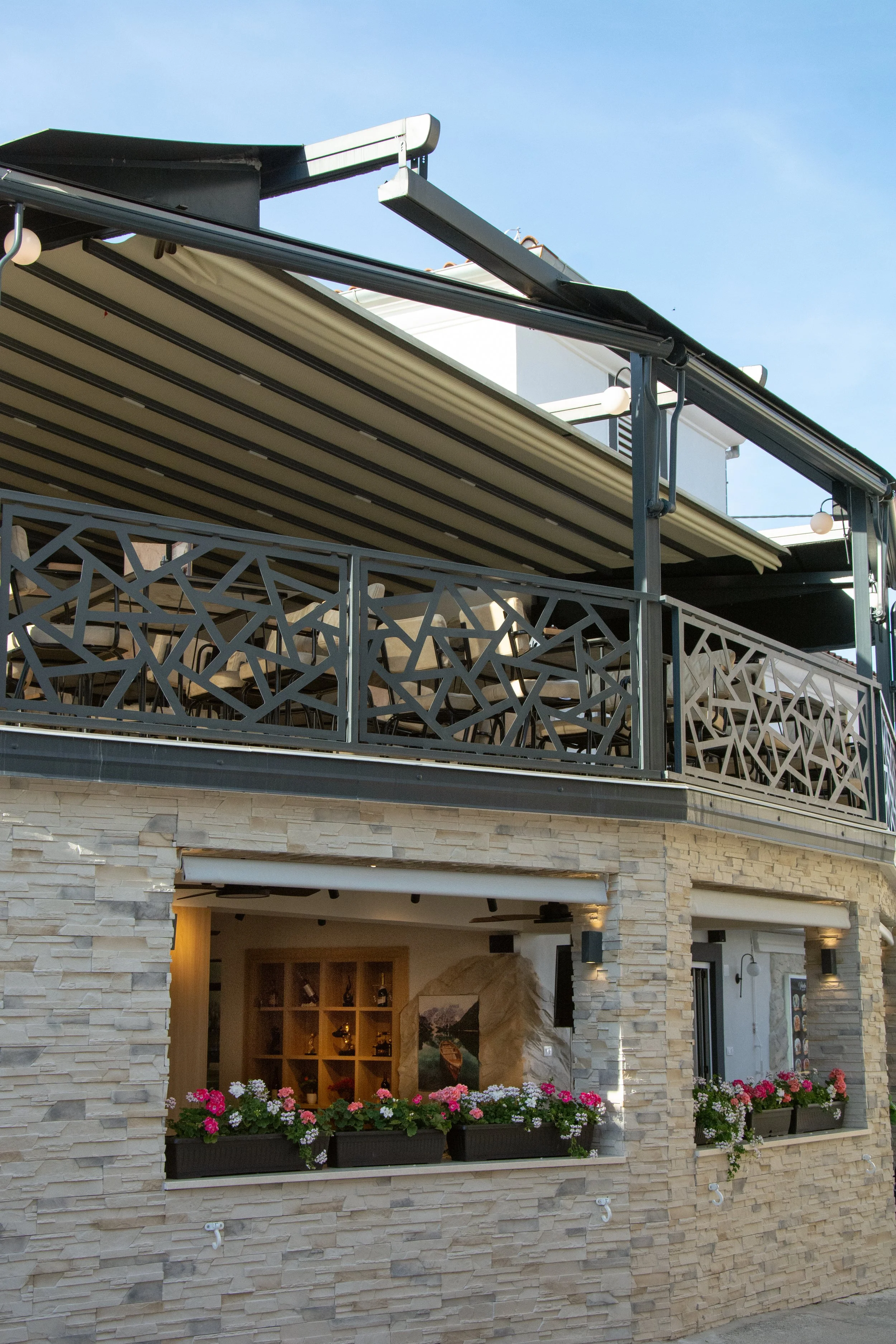 Balcony with flower boxes filled with pink and white flowers, behind a decorative metal railing, part of a building with stone facade, and a blue sky in the background.