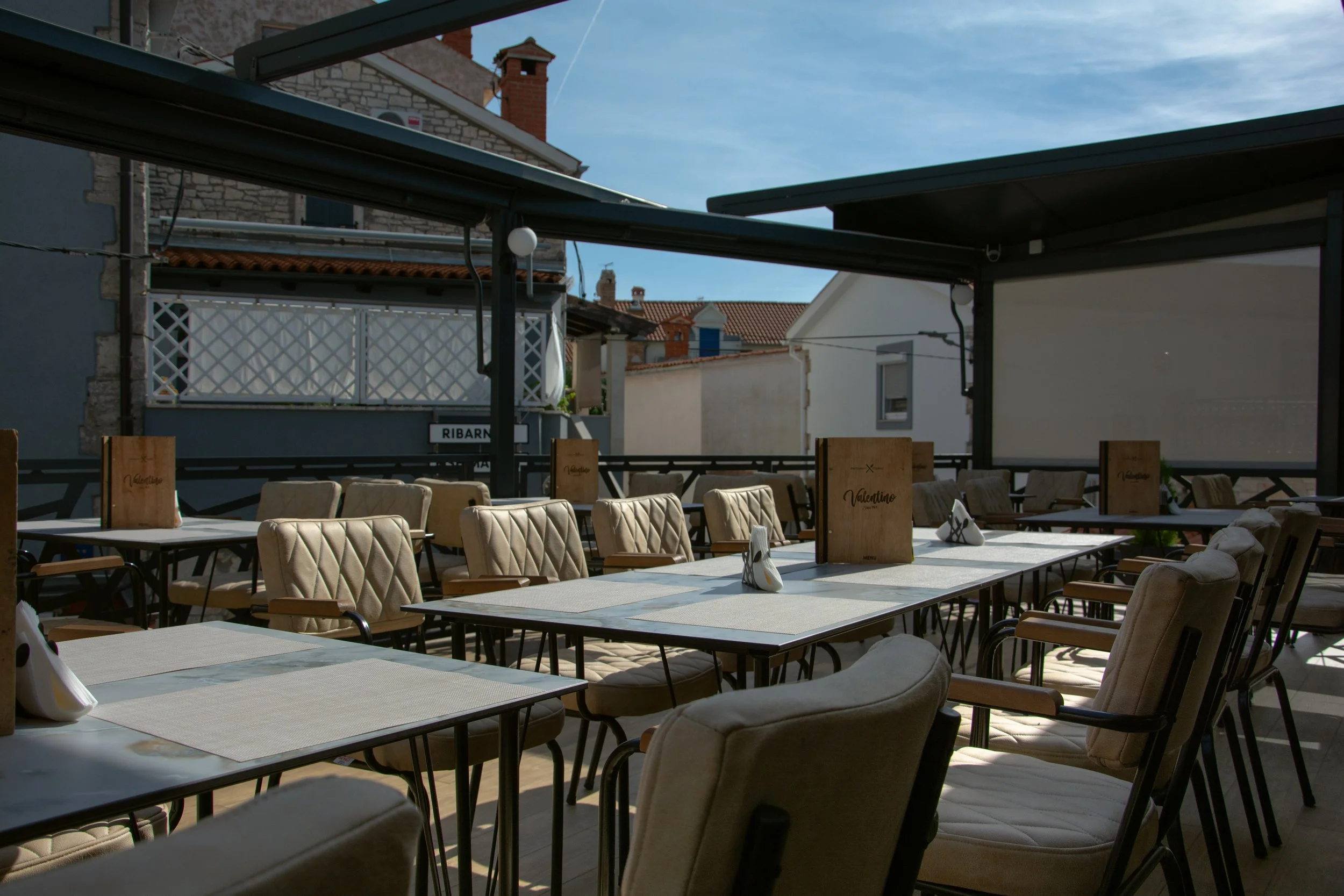 Empty outdoor restaurant or cafe terrace with tables and cushioned chairs, under a partially covered area, with buildings and a blue sky in the background.