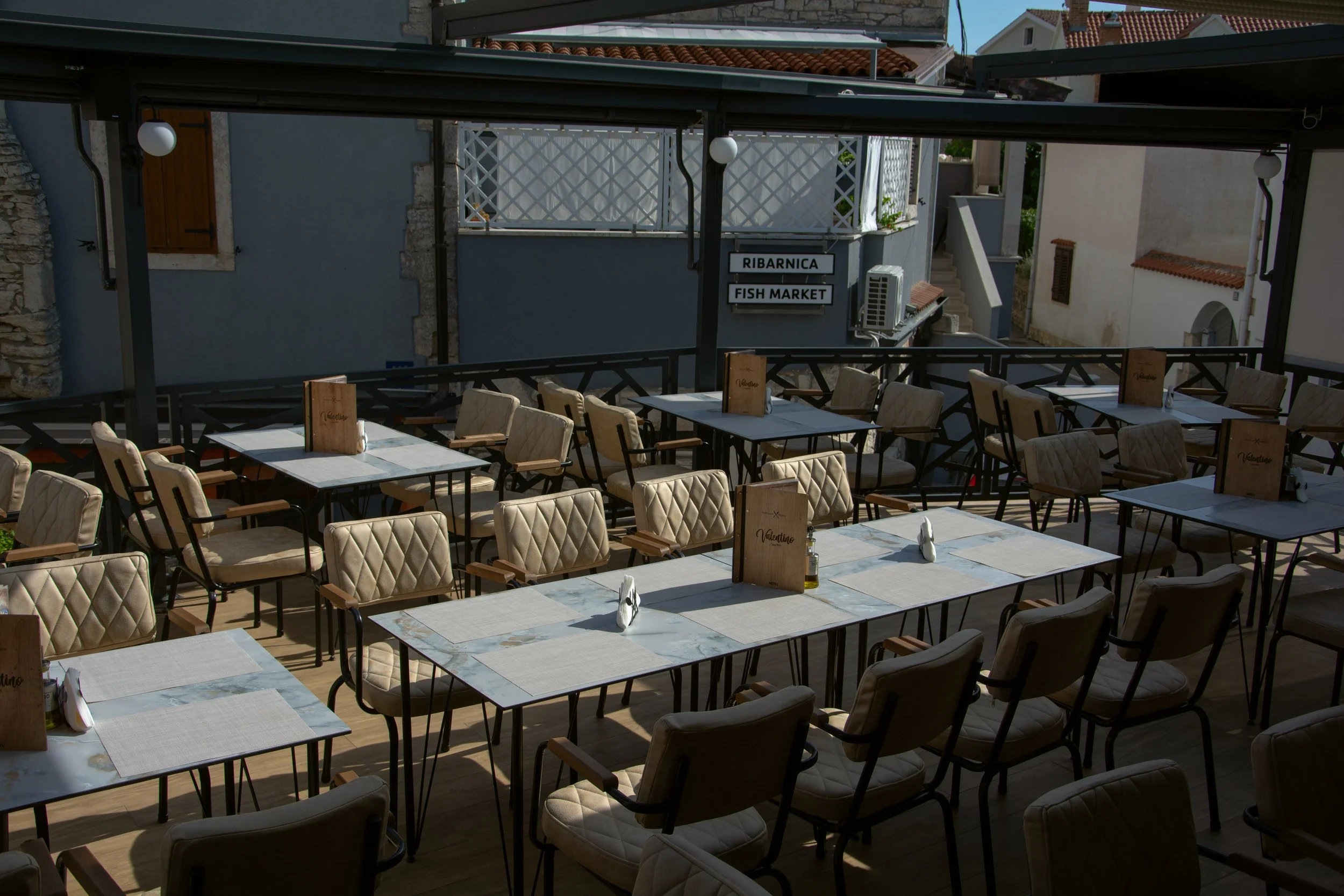 Empty outdoor restaurant patio with tables and chairs, signs reading 'Ribarnica' and 'Fish Market', and a decorative railing, under a covered structure.