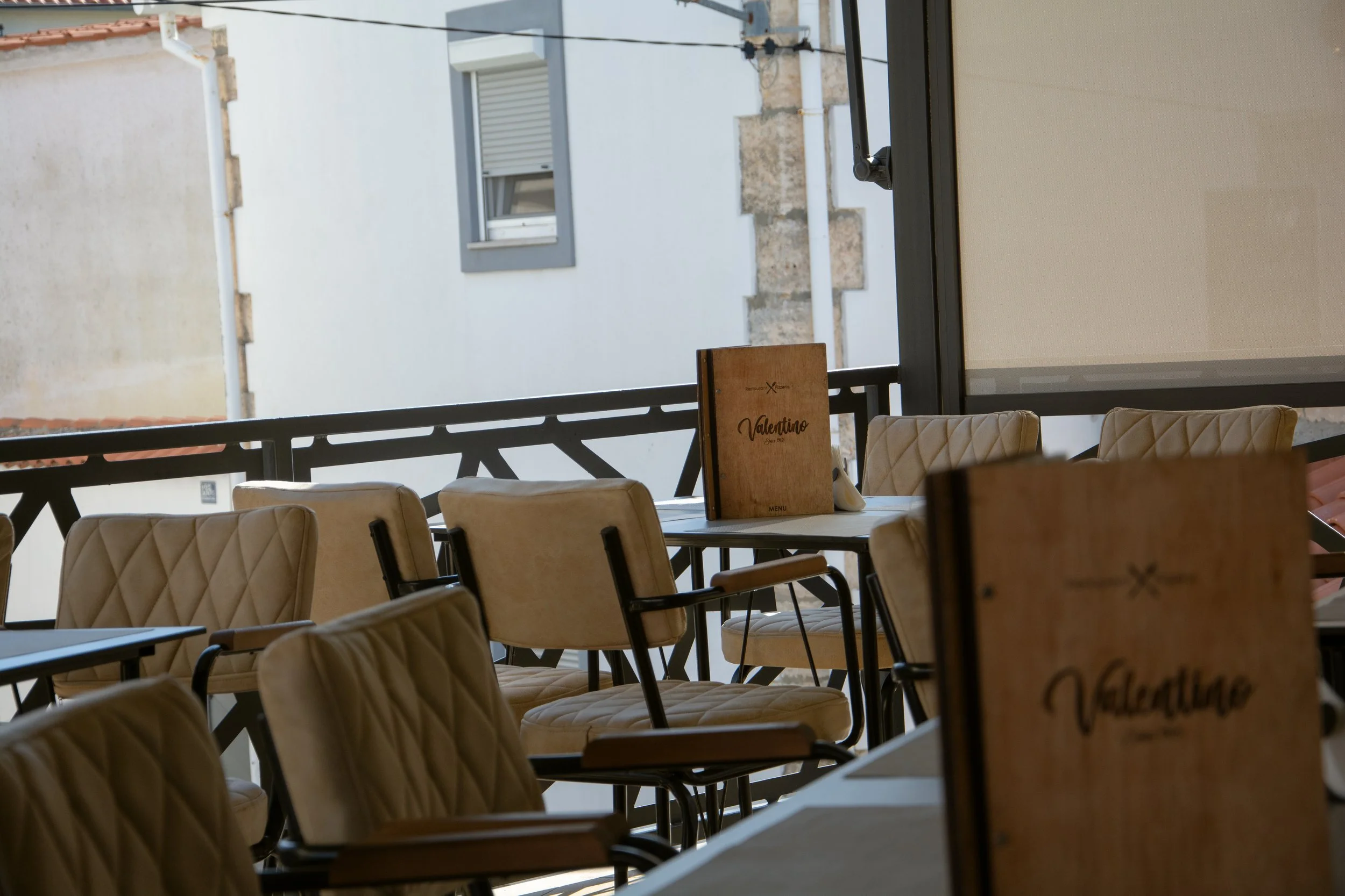 Restaurant interior with beige cushioned chairs around tables, a wooden menu on one table, and a large window showing buildings outside.