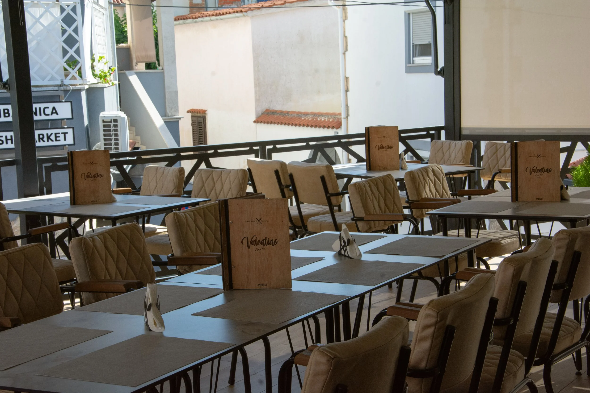 Empty outdoor restaurant seating area with beige cushioned chairs and tables set with placemats and menus, overlooking neighboring buildings with white walls and red tile roofs.