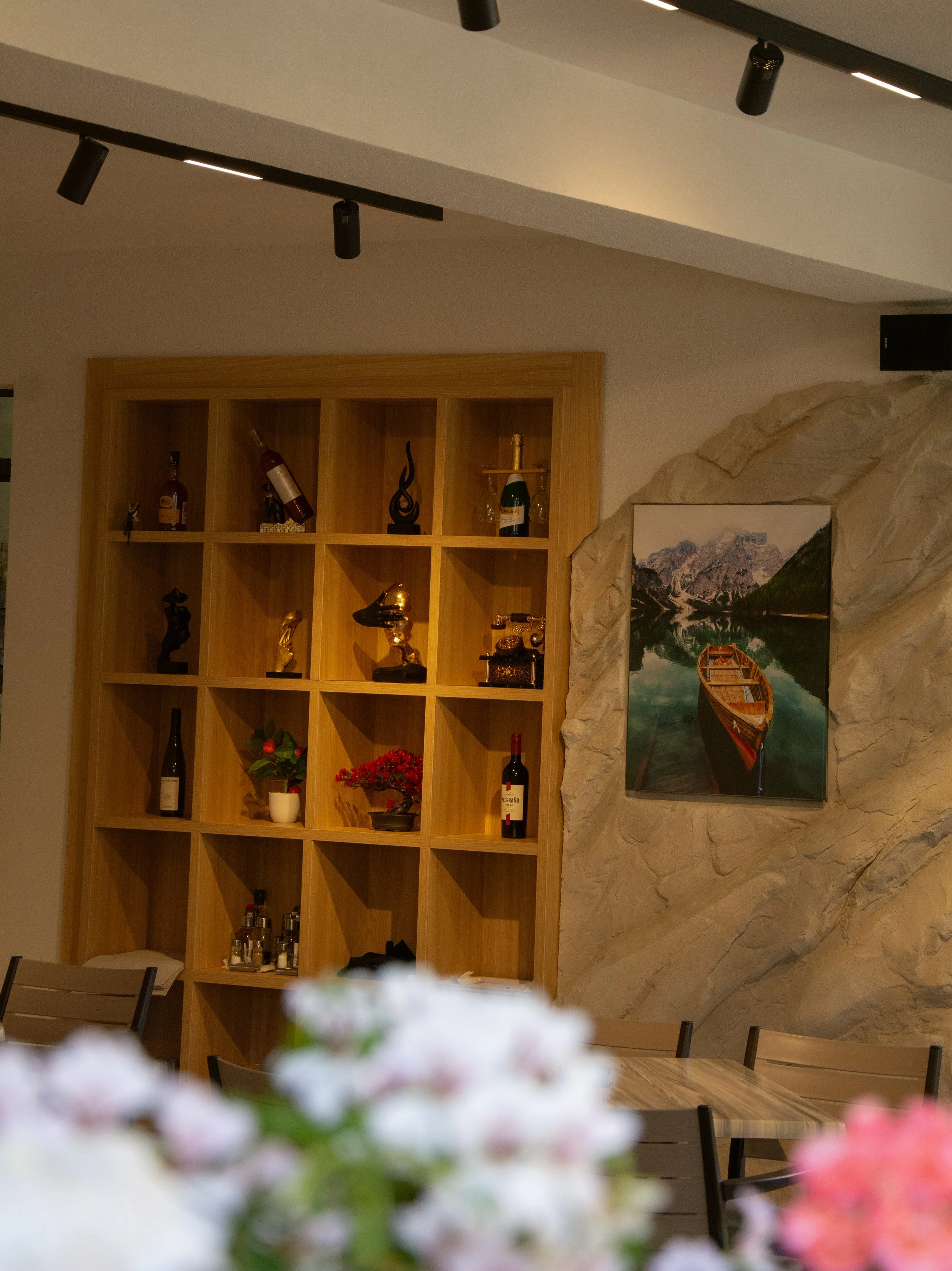 Interior of a room featuring a large wooden shelf with decorative bottles and sculptures, a framed landscape photograph of mountains and a lake, and pink and white flowers in the foreground.