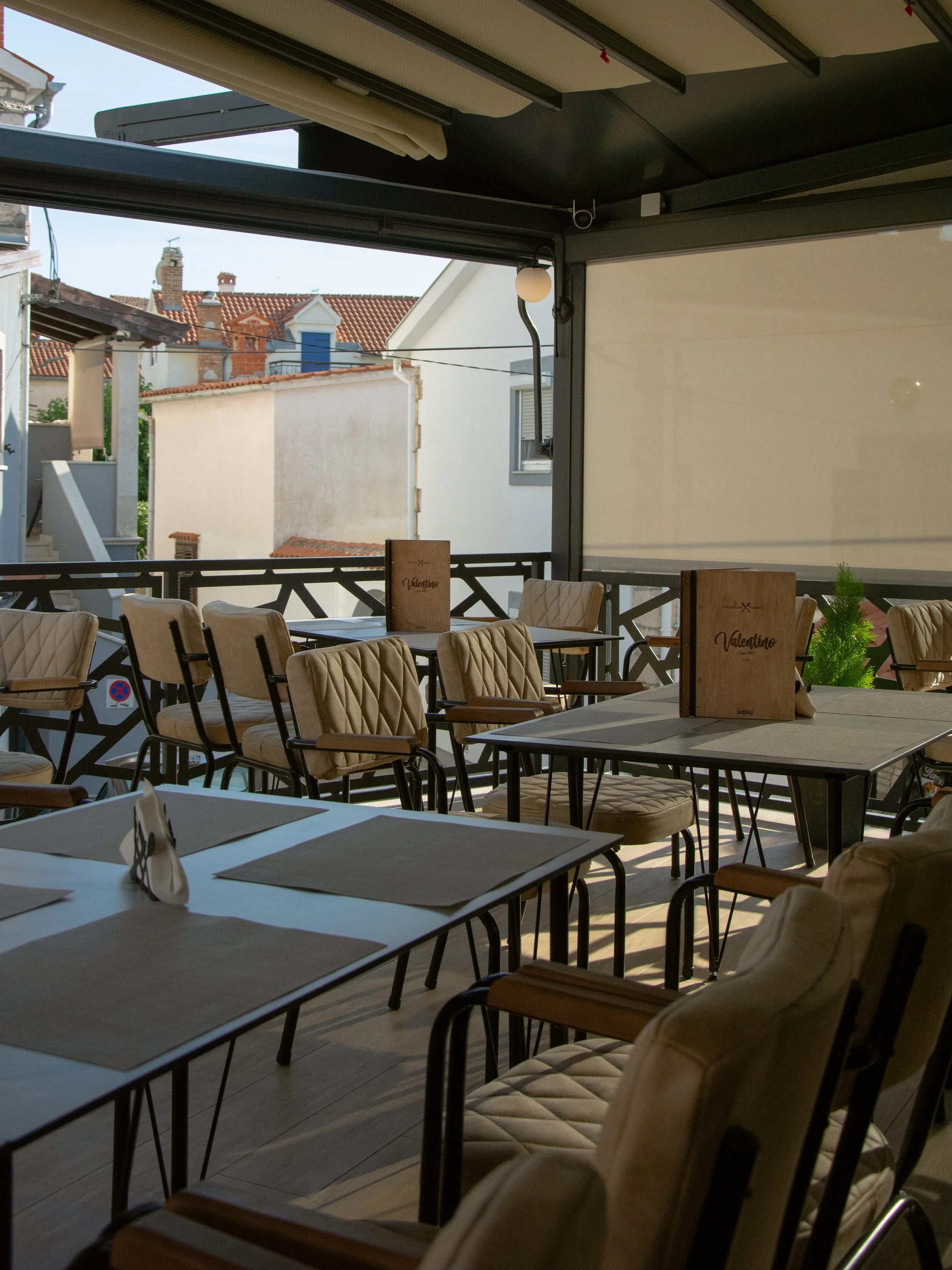 An outdoor dining area with several beige cushioned chairs and tables set with placemats, umbrellas, and a menu standing upright on the table. The background shows neighboring rooftops and buildings under a clear sky.