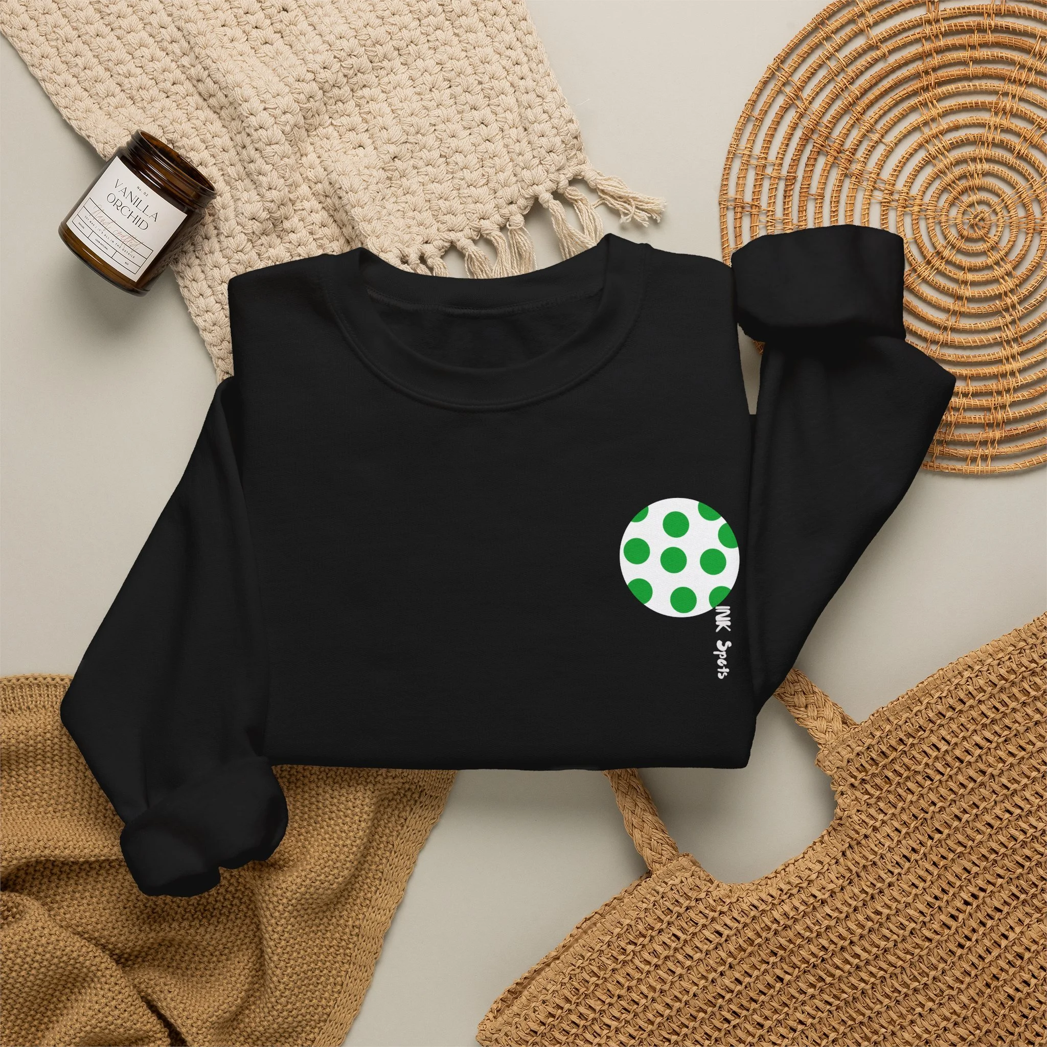Flat lay of various summer accessories including a black t-shirt with a green polka dot design, a beige crochet hat, a tan woven handbag, a brown wicker placemat, a cream textured scarf, and a candle labeled vanilla orchid.