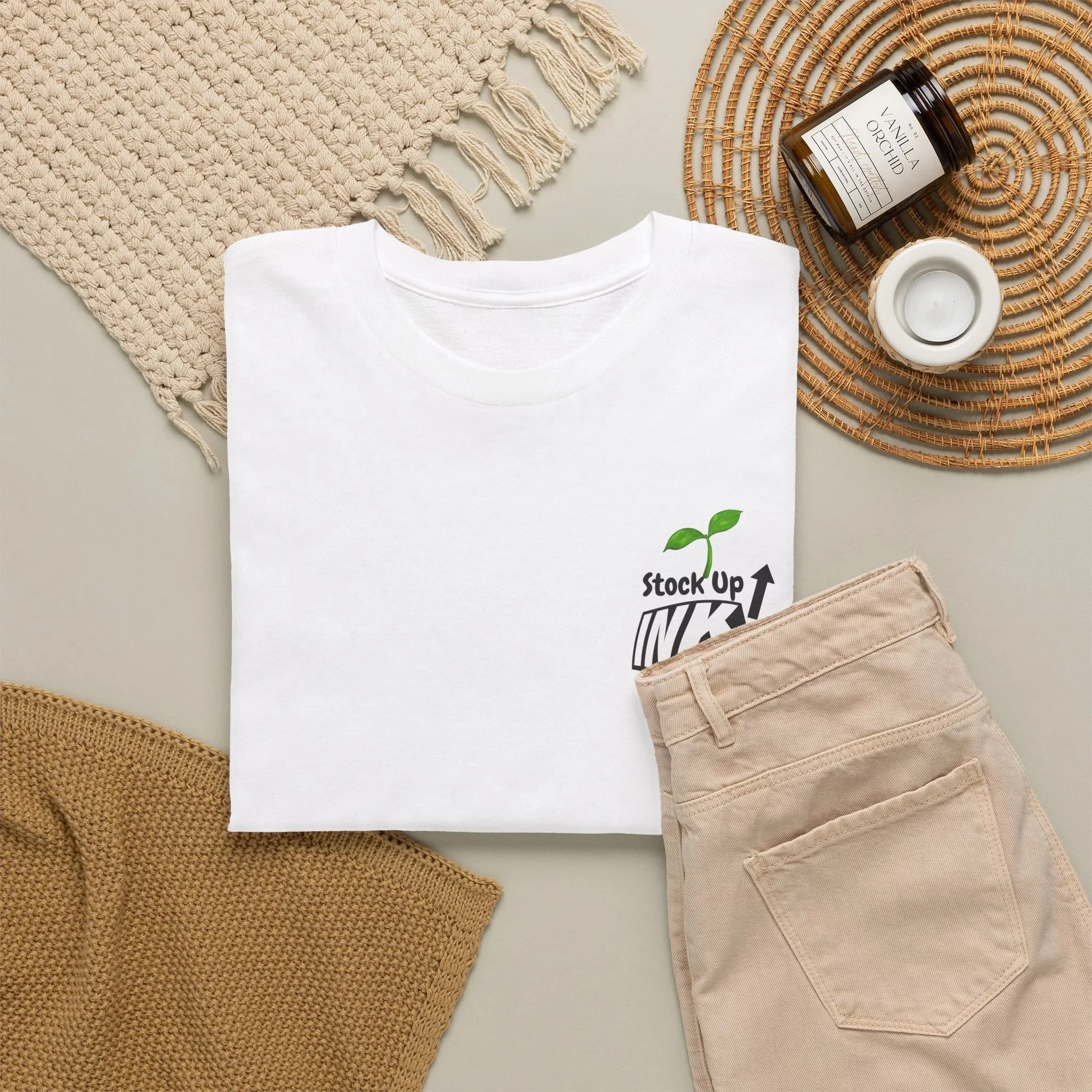 A flat lay of neutral-colored clothing and decor items including a white T-shirt, beige pants, a knitted brown piece, a woven beige bag, a beige woven hat, a cream-colored fringed cloth, a dark amber glass jar labeled "Vanilla Orchid," a small white candle, and a round rattan placemat.